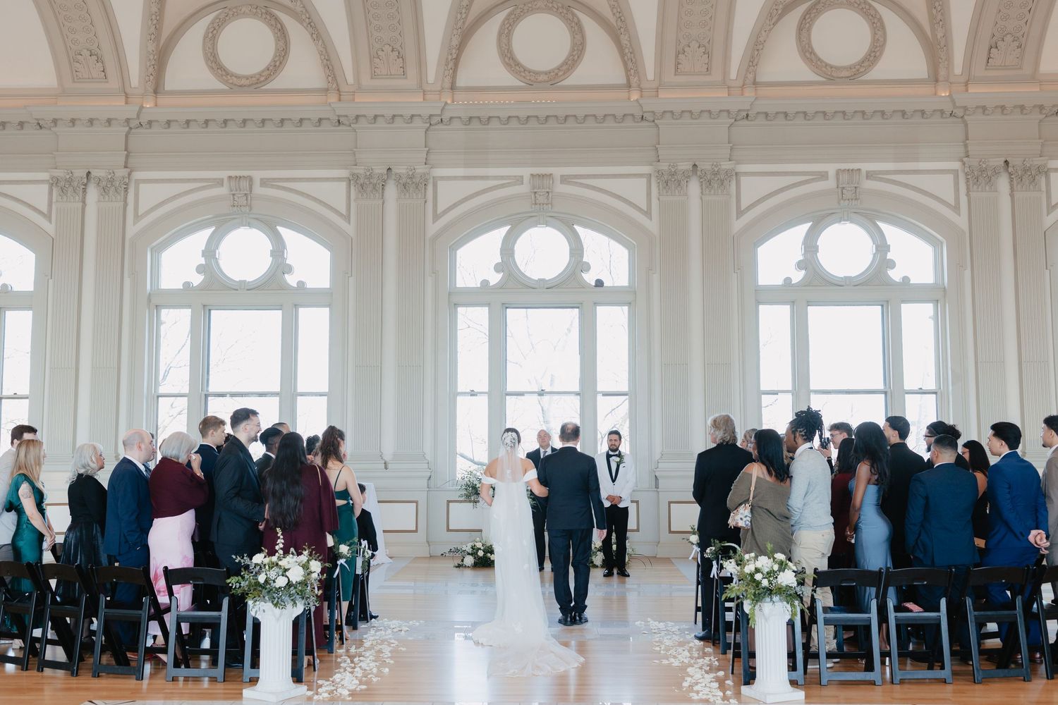 Elegant wedding ceremony in grand ballroom with tall arched windows and white floral arrangements.
