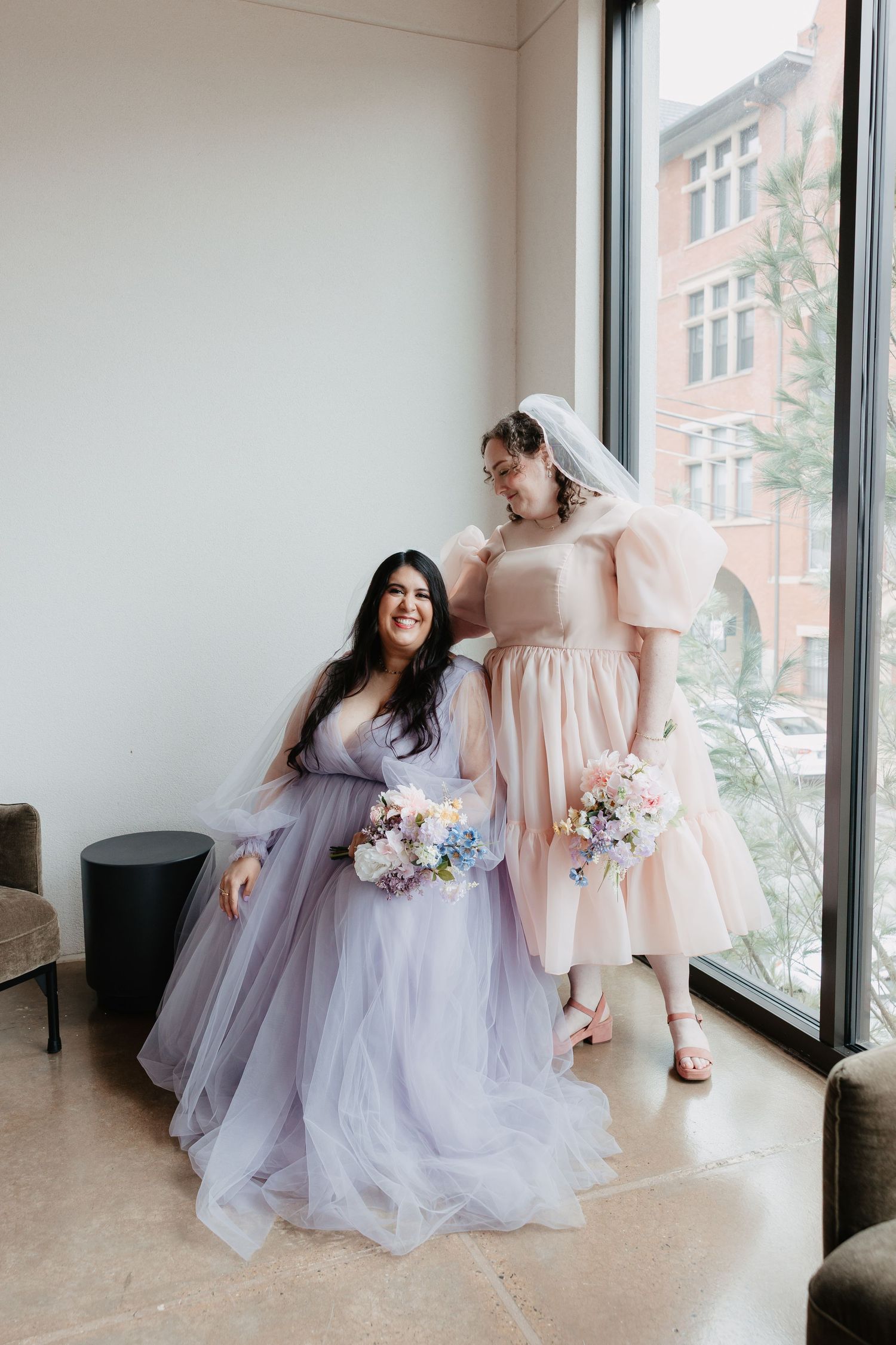 Two brides pose by large windows, one in lavender tulle gown, one in blush pink dress holding bouquets.