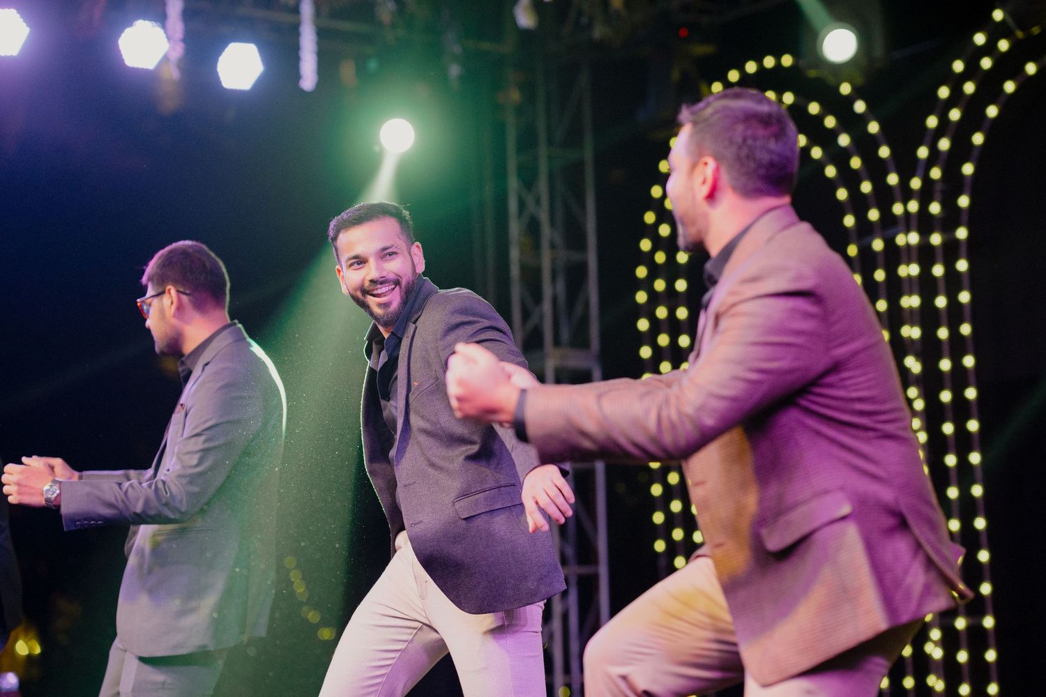 Three men in suits dancing joyfully on a lit stage at an outdoor evening event.