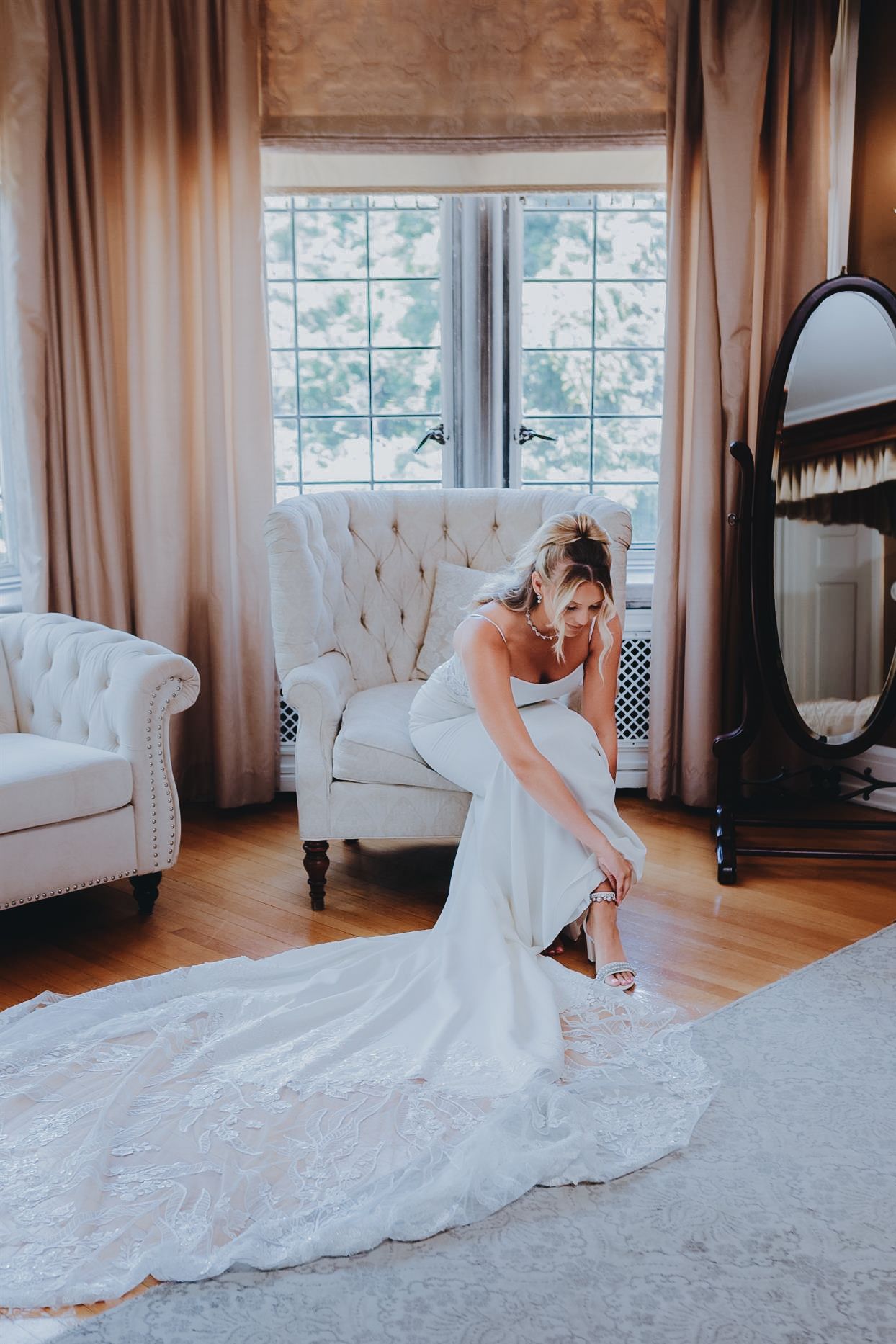 Bride in elegant white gown adjusting her shoe beside a tufted white chair in a beautifully decorated bridal suite. In Philadelphia PA