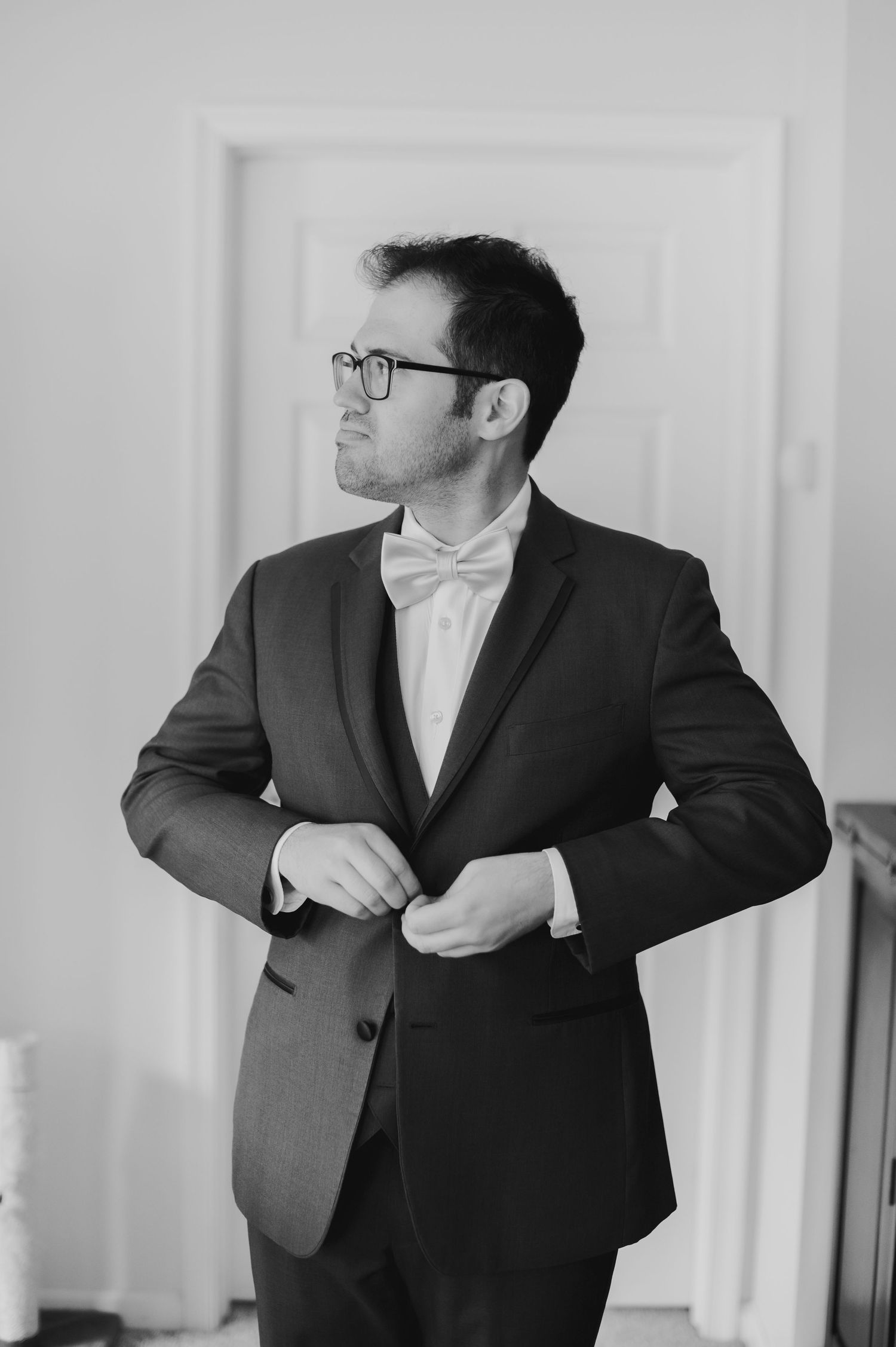 Black and white photo of a groom in a suit and bow tie buttoning his jacket before the wedding ceremony.