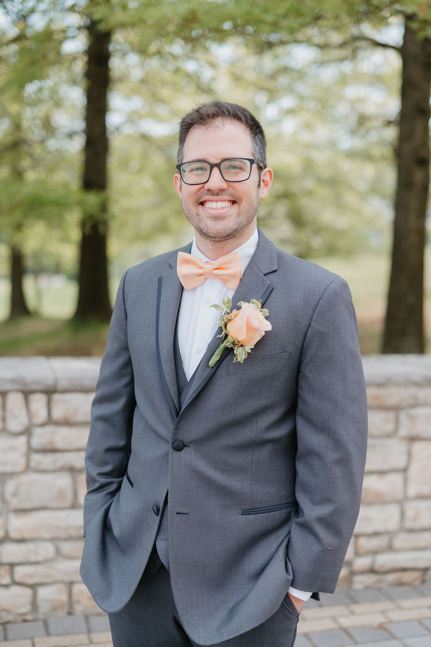 Smiling groom in gray suit with peach bow tie and boutonniere posing outdoors near stone wall and trees.