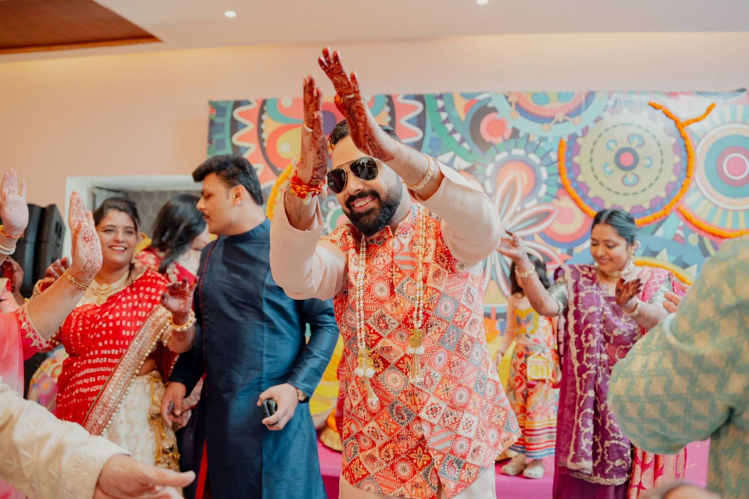 Groom in orange floral garland and sunglasses celebrates with raised hands at vibrant Indian wedding ceremony in Philadelphia PA