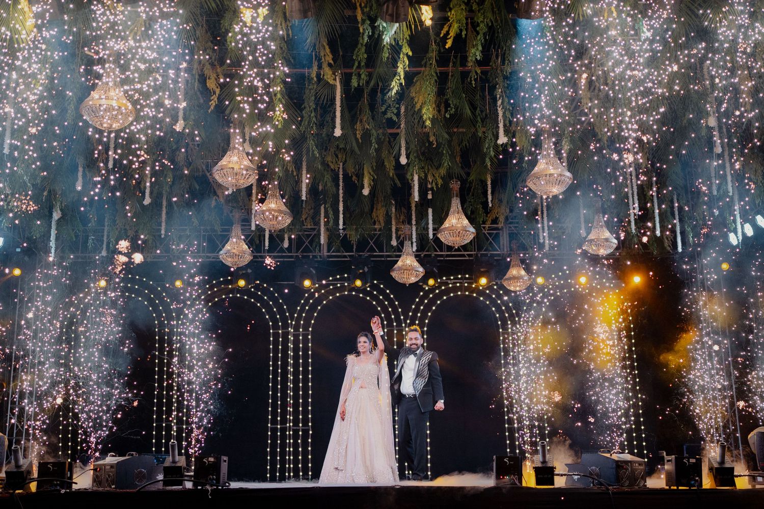 Bride and groom stand on grand stage with sparkling fireworks, crystal chandeliers, and lush greenery at elegant wedding.