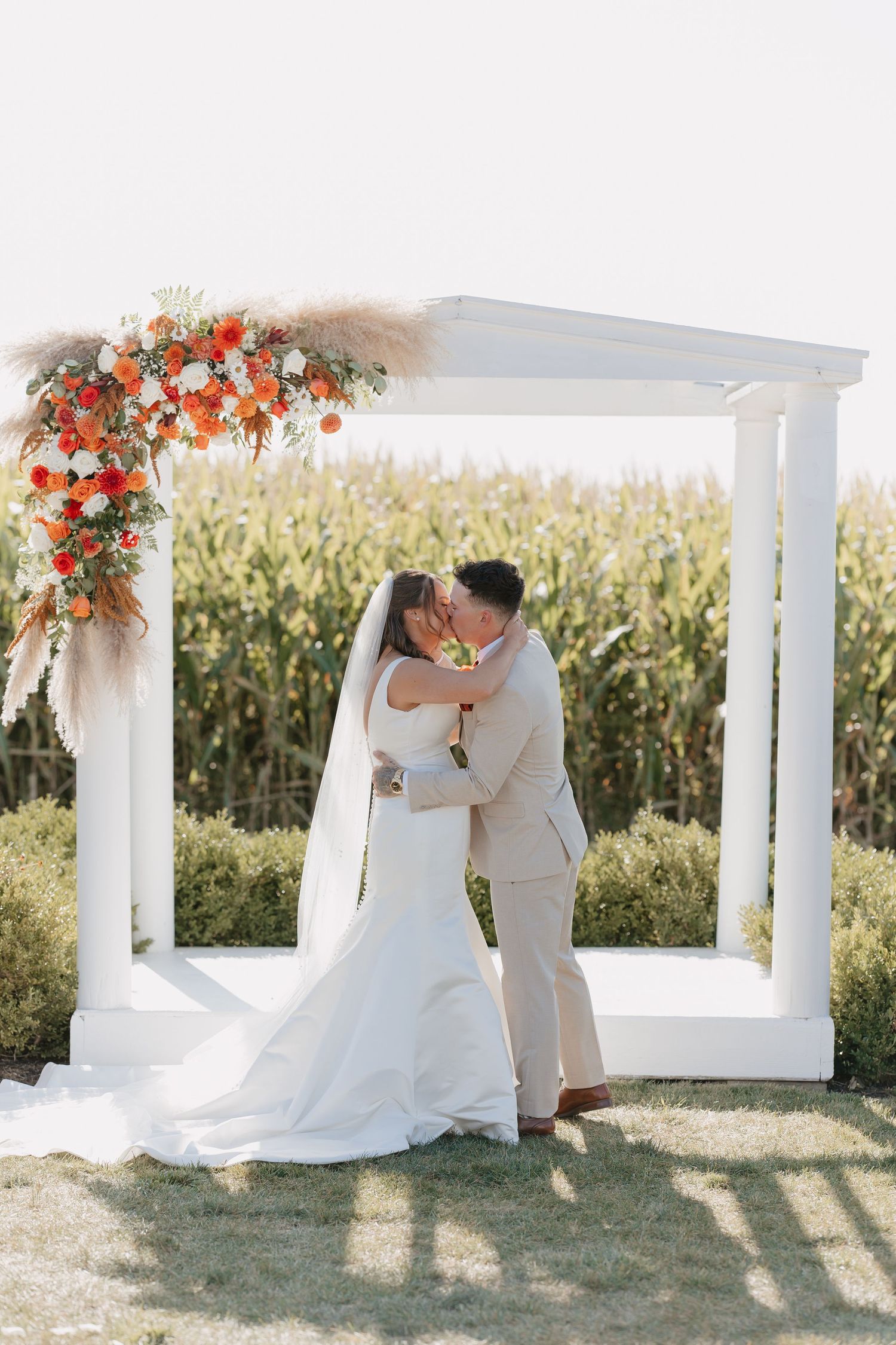 Bride and groom share a first kiss under a white floral arch with orange flowers beside a cornfield.