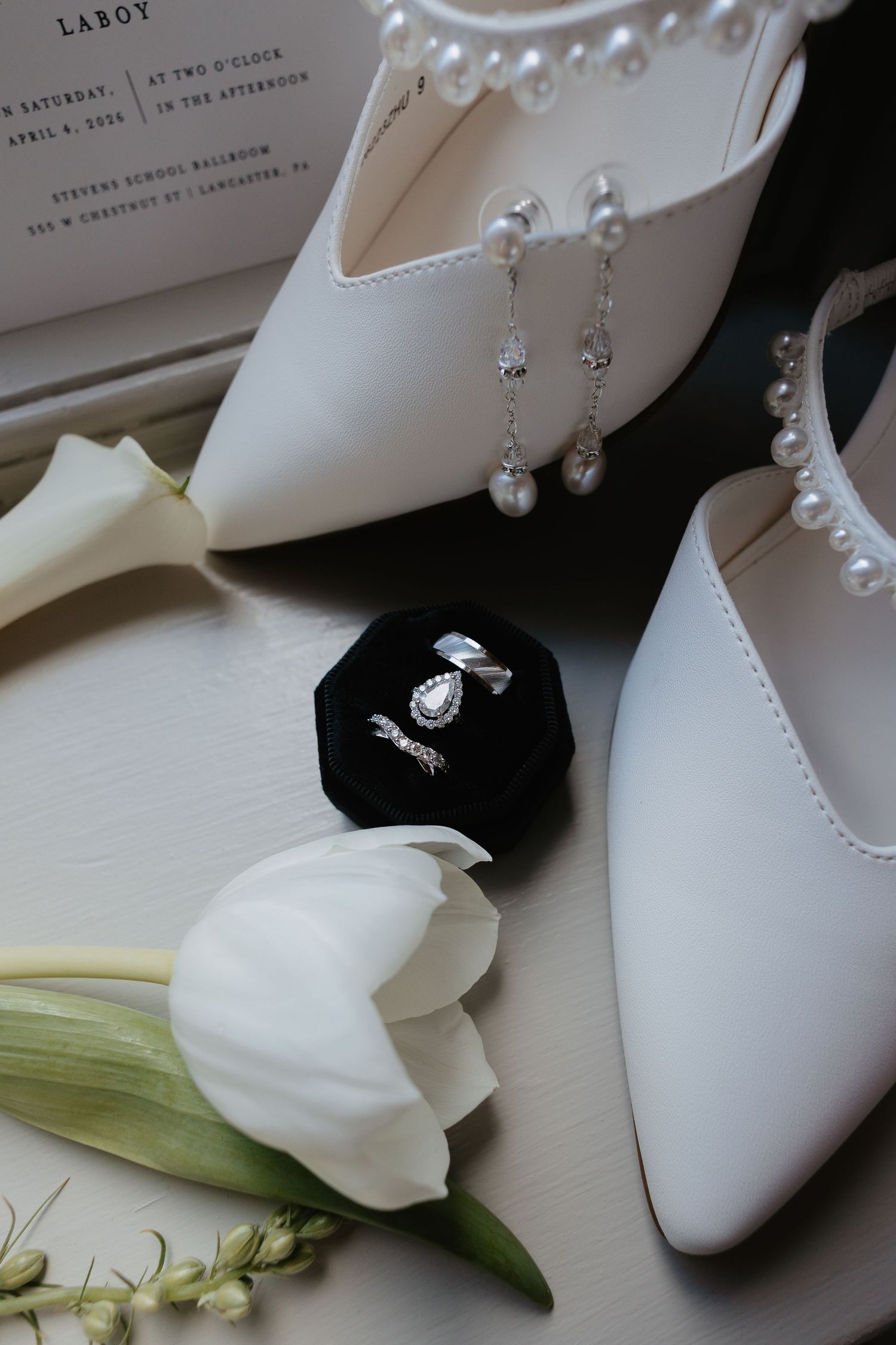 Bridal accessories flatlay with pearl shoes, diamond rings in velvet box, and white tulip.