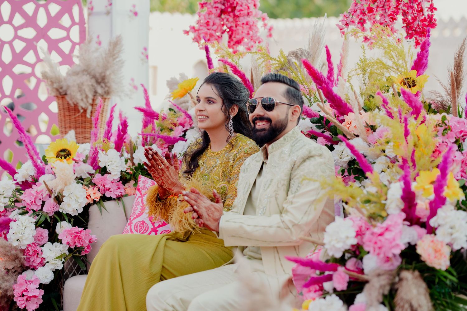 Couple in traditional Indian wedding attire seated among vibrant pink and yellow floral decorations at a festive ceremony.