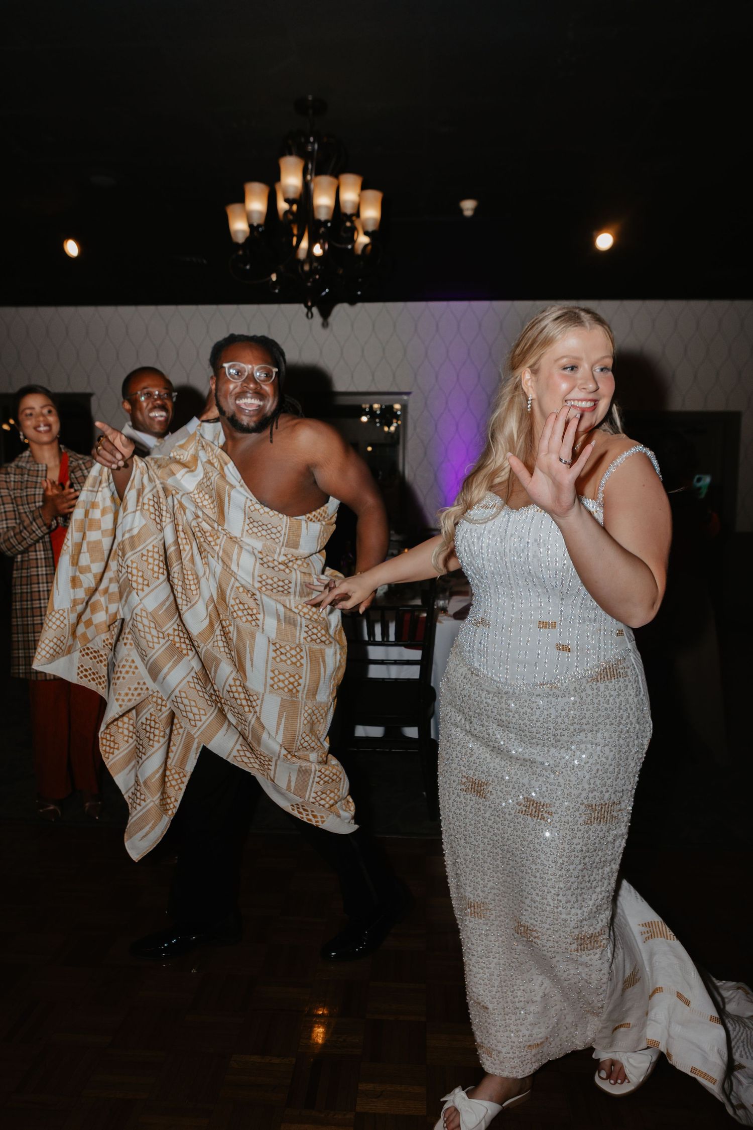 A bride in a sequined gown dances joyfully with a guest draped in a patterned cloth at a wedding reception at a Traditional Nigerian Wedding
