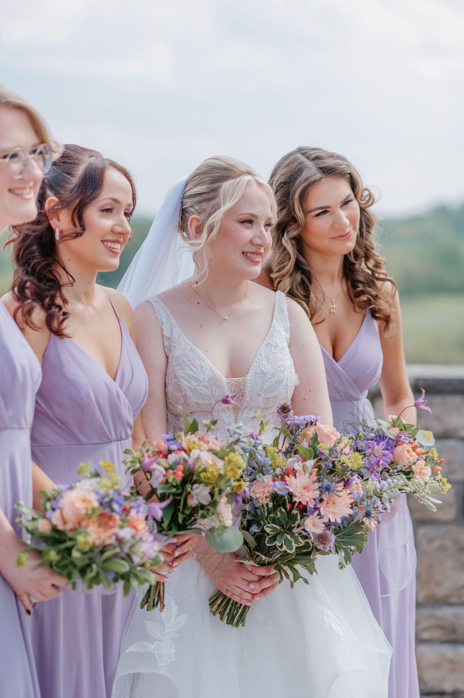 Bride in white lace gown with bridesmaids in lavender dresses holding colorful floral bouquets outdoors. The View at Morgan Hill Wedding