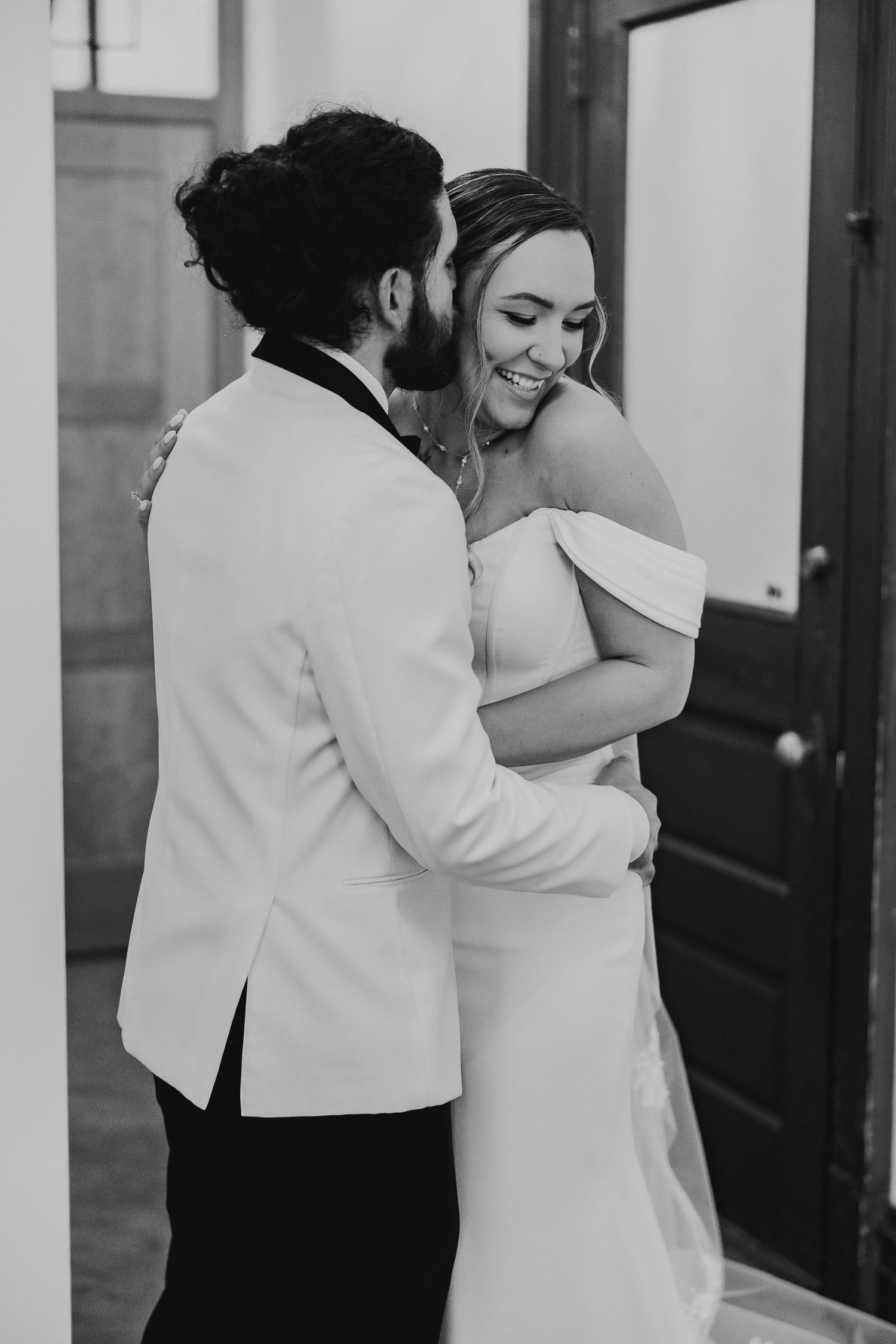 Groom whispering to laughing bride in off-shoulder gown, romantic black and white wedding portrait.