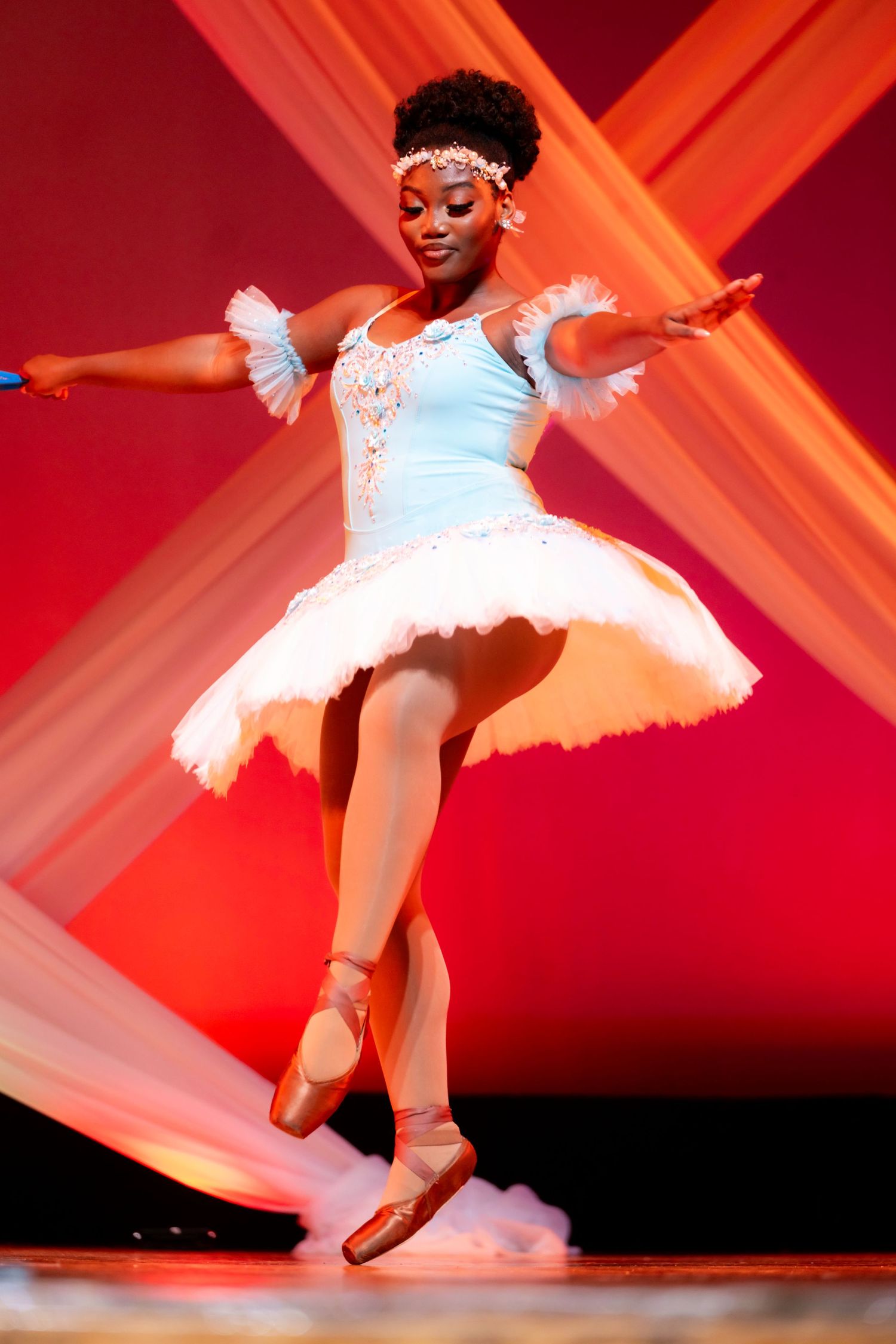 Young Black ballerina in white tutu and pointe shoes performs graceful pirouette on stage with red backdrop.