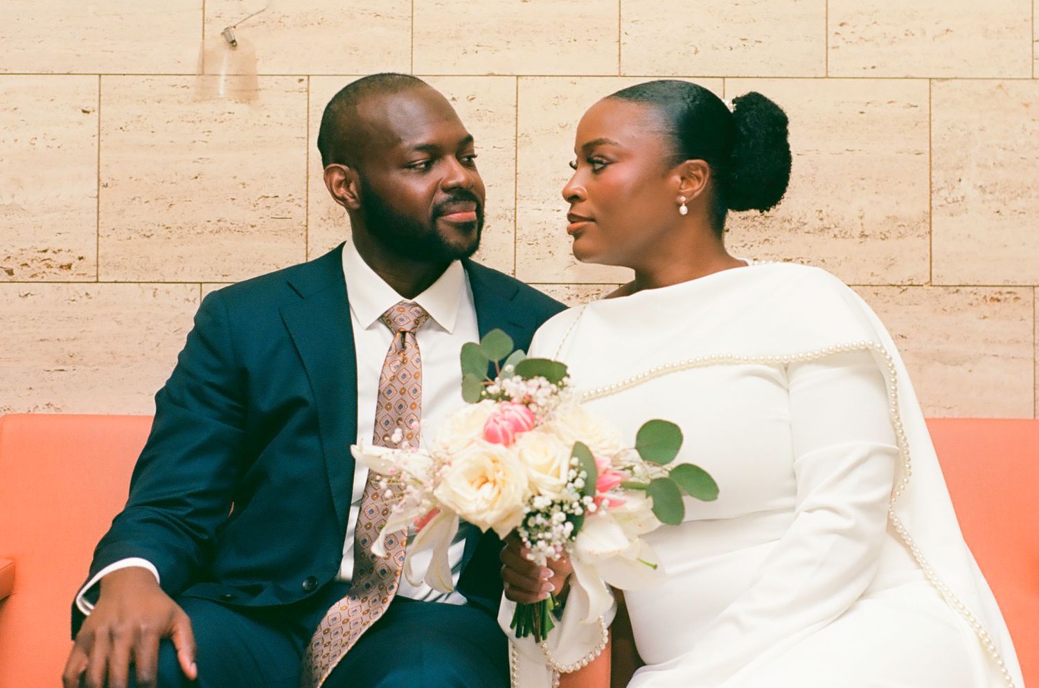 A newlywed couple in wedding attire sits together, the bride holding a pink bouquet, gazing lovingly at each other.