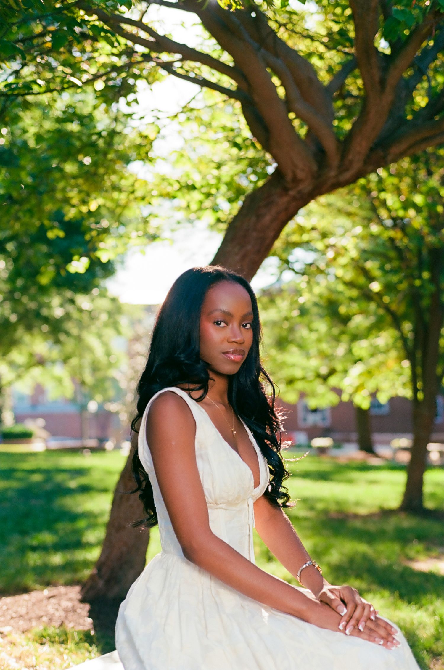 A young Black woman in a white dress sits gracefully under a lush green tree in a sunlit park.