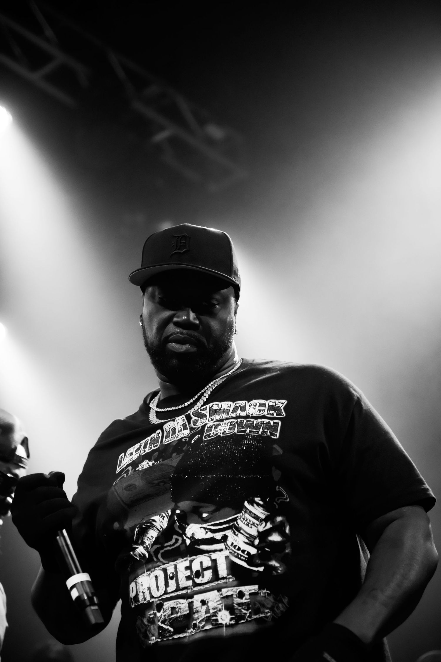 Black and white photo of a rapper performing on stage holding a microphone wearing a cap and graphic tee.