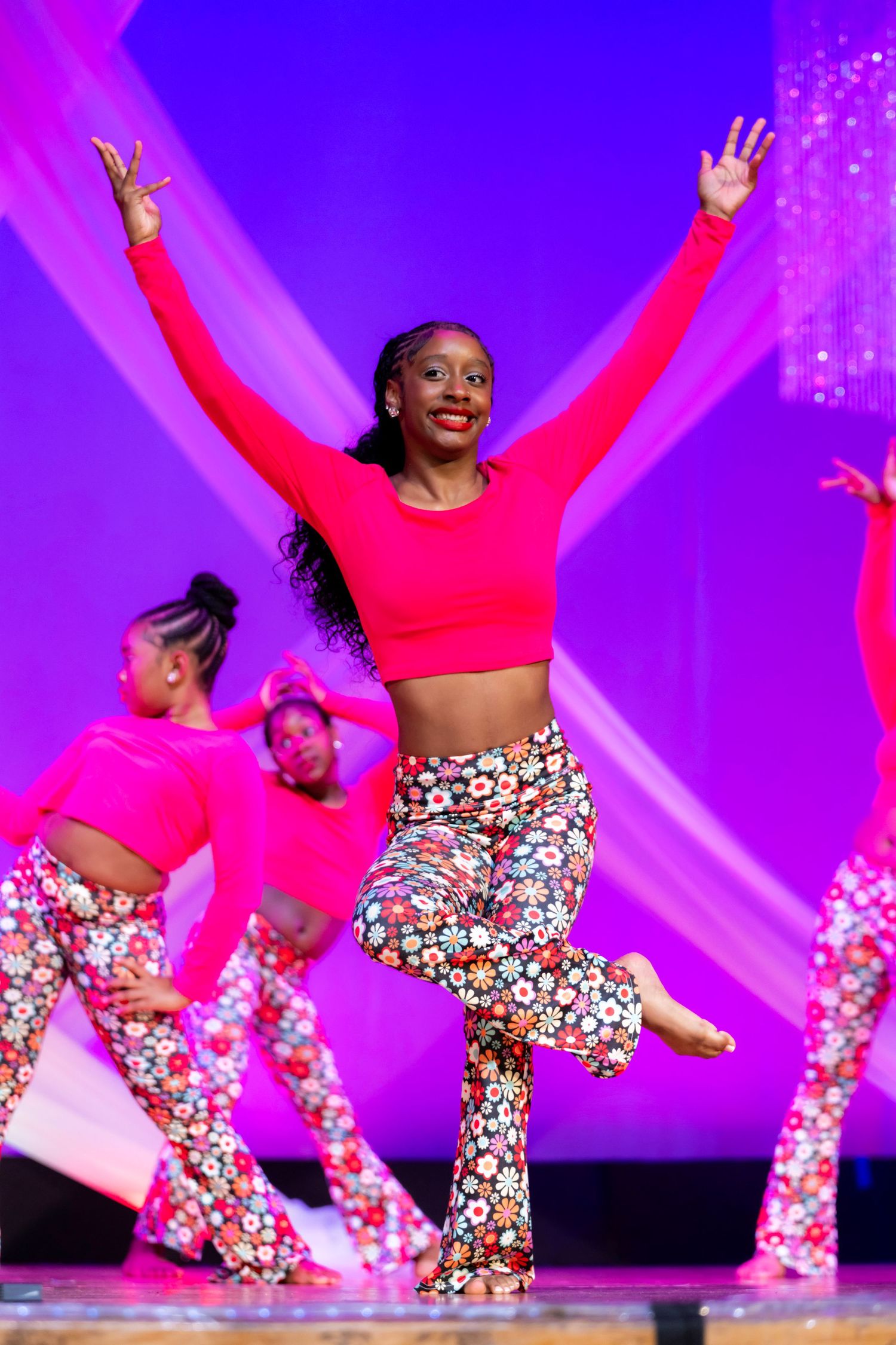 Young Black dancer in red crop top and floral pants performs on stage with arms raised, backed by dancers in matching outfits.