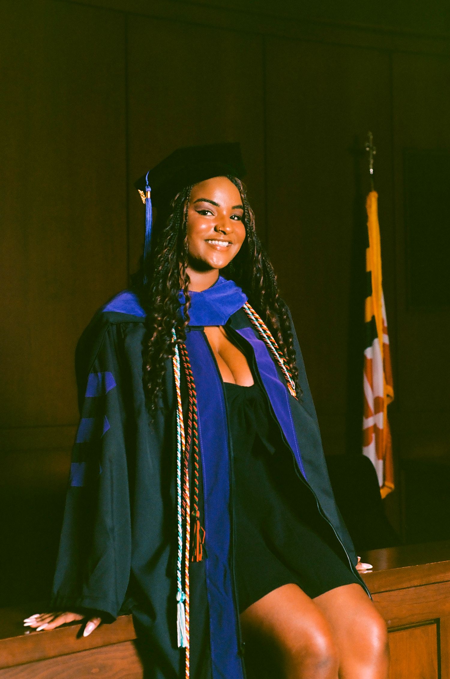 A smiling graduate in black and blue cap and gown with honor cords poses proudly near a flag indoors.