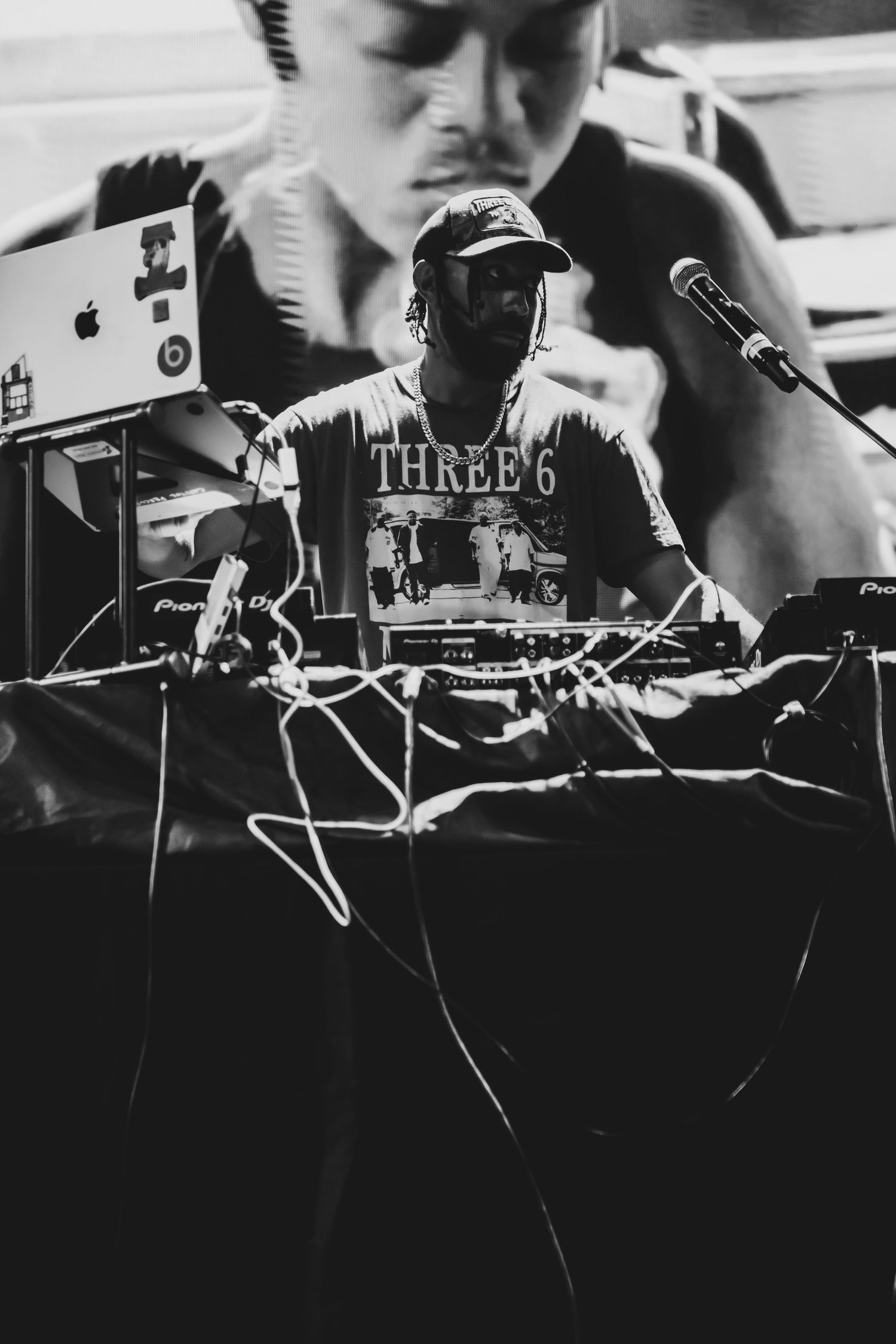 Black and white photo of a DJ wearing a Three 6 Mafia shirt performing with Pioneer DJ equipment and a laptop on stage.