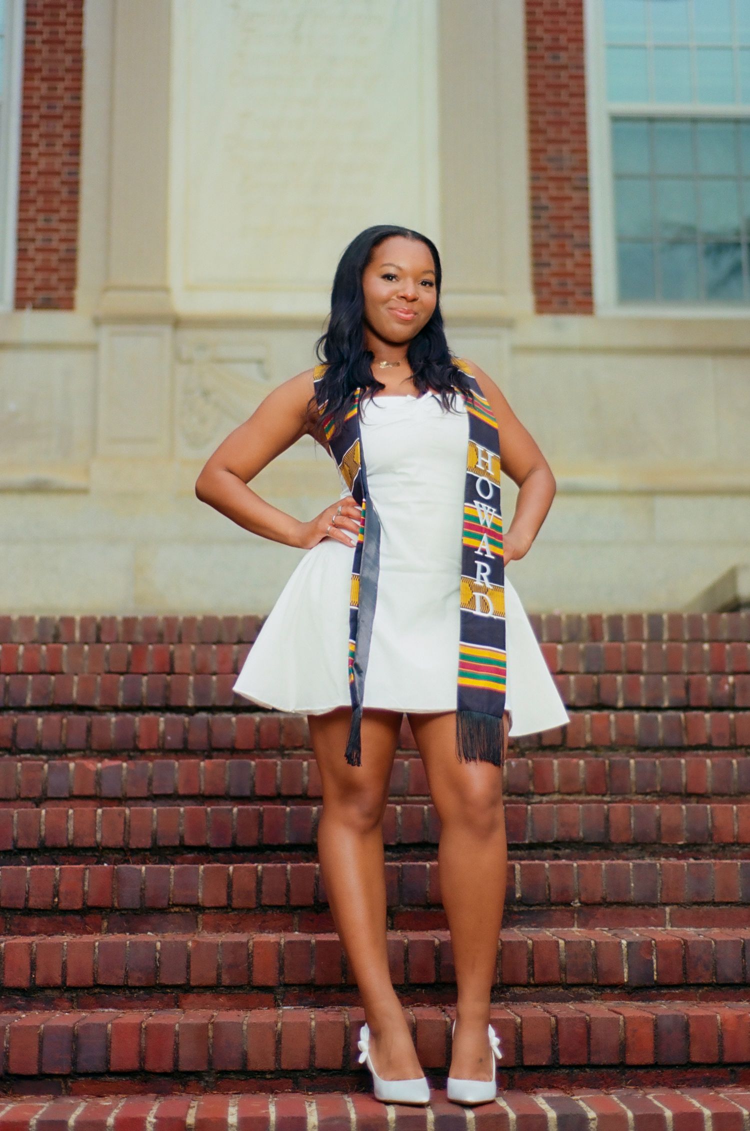 A young woman in a white dress and Howard University kente graduation stole poses proudly on brick steps.