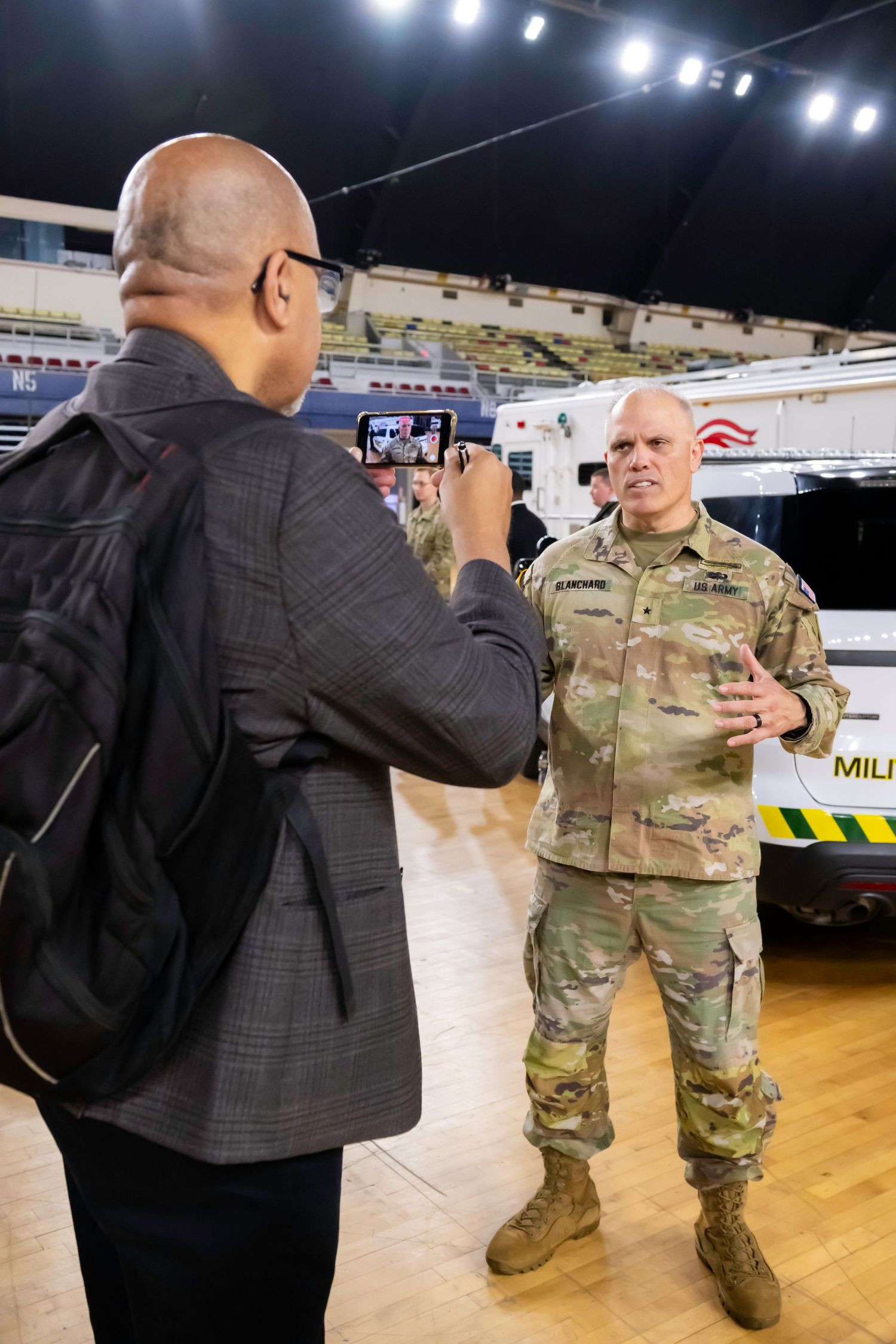 A civilian photographs a U.S. Army soldier in camouflage uniform inside a large indoor arena.