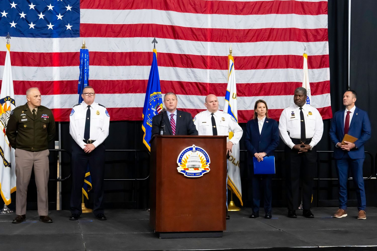 Law enforcement officials stand at podium before American flag at official press conference.