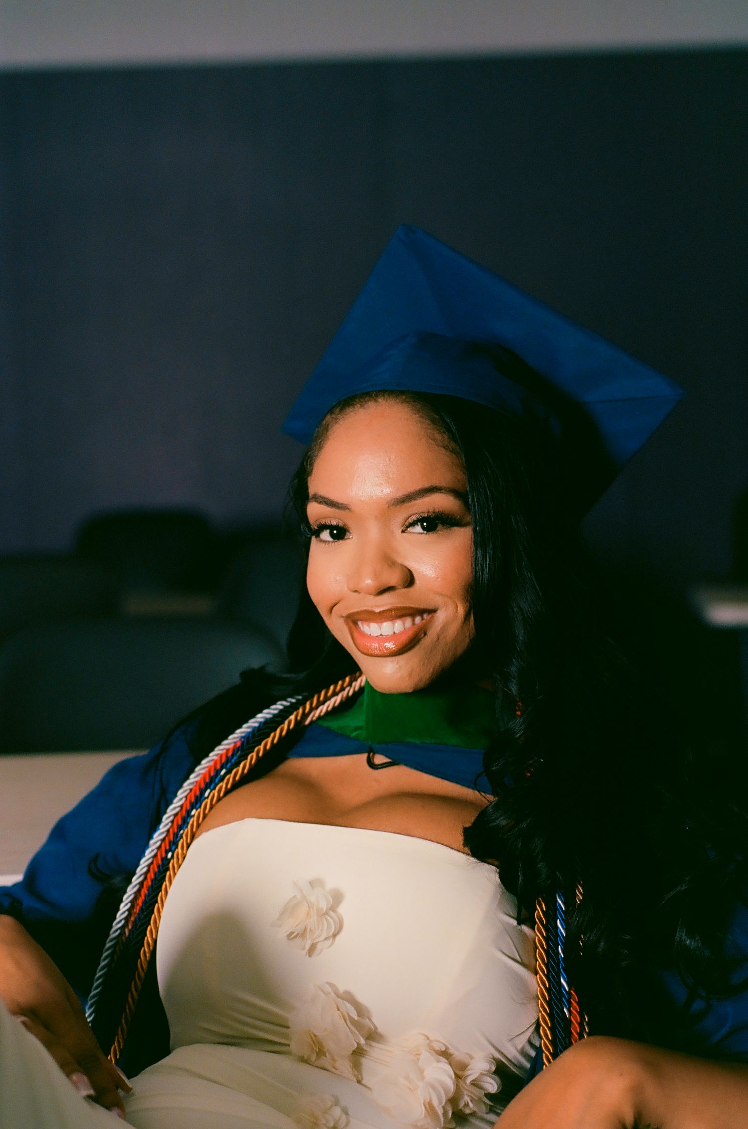 Smiling graduate in blue cap and gown with honor cords poses for a portrait photo at graduation ceremony.