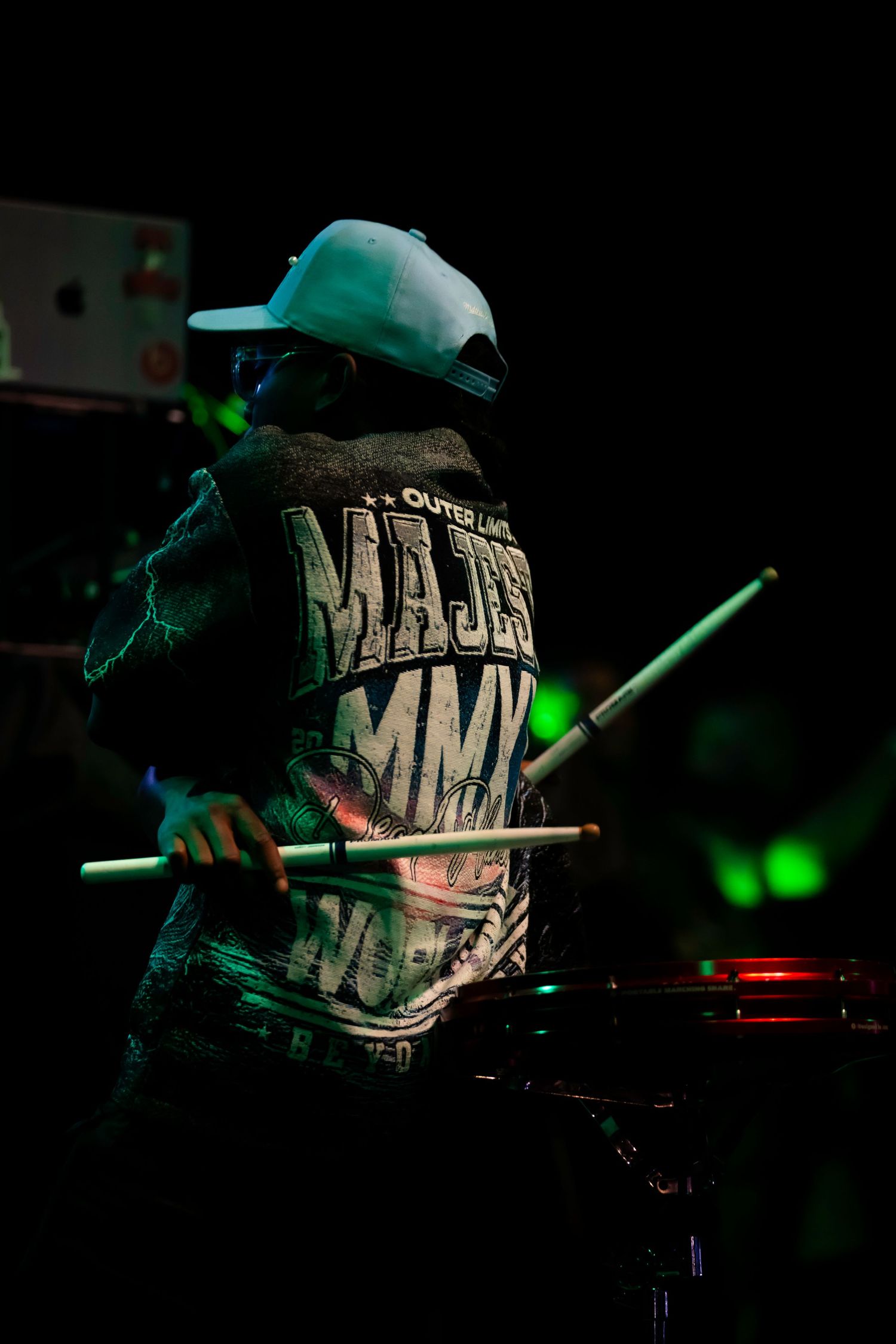 Drummer performing on stage in green stage lighting, wearing a graphic hoodie and white cap, holding drumsticks.