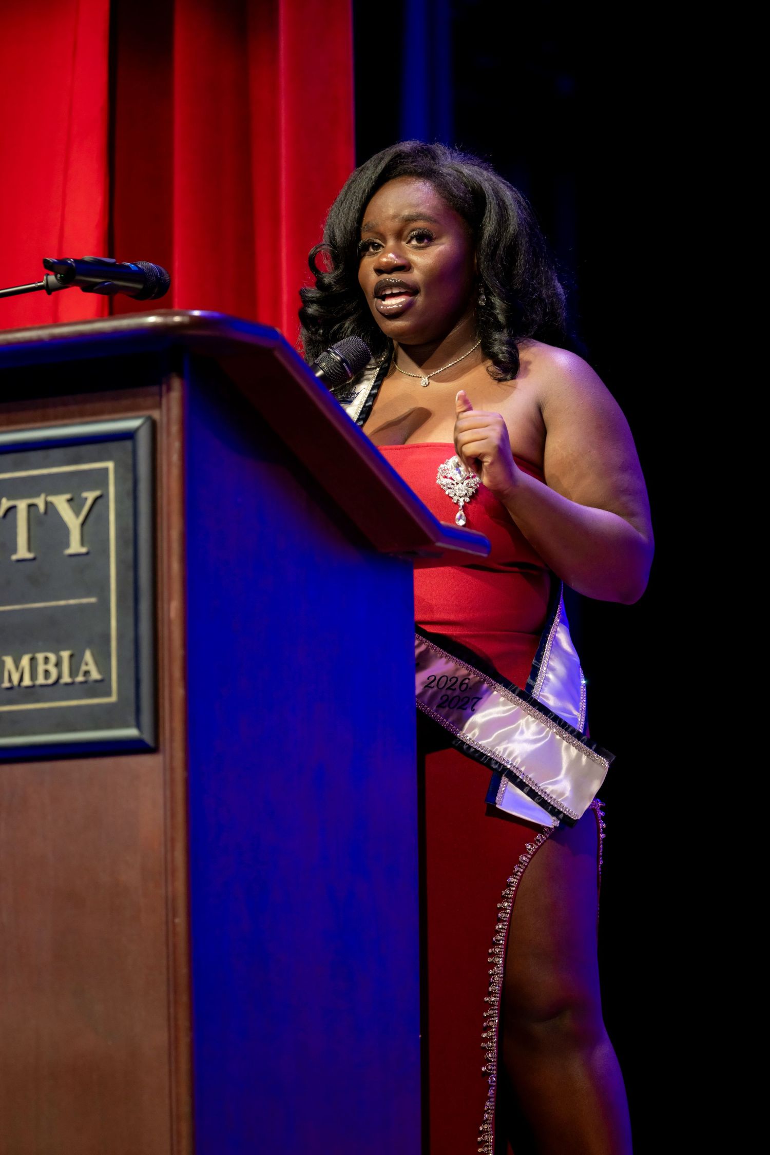 Woman in red gown and sash speaks at university podium on stage with red curtain backdrop.
