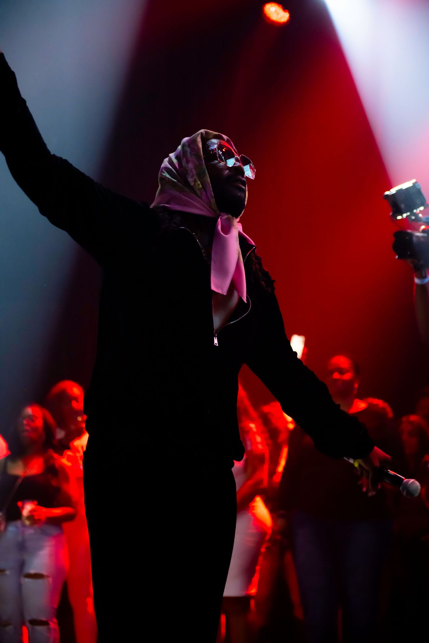Performer on stage raising one hand wearing a headscarf and sunglasses under dramatic red stage lighting.