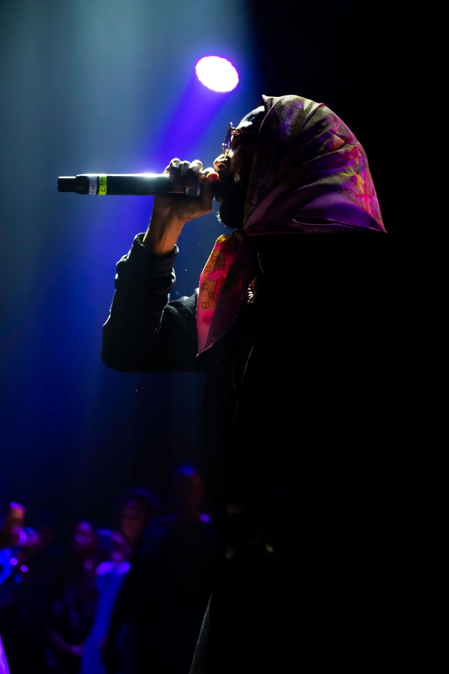 Silhouetted performer singing into microphone on stage, wearing colorful headscarf, illuminated by blue and purple concert lights.