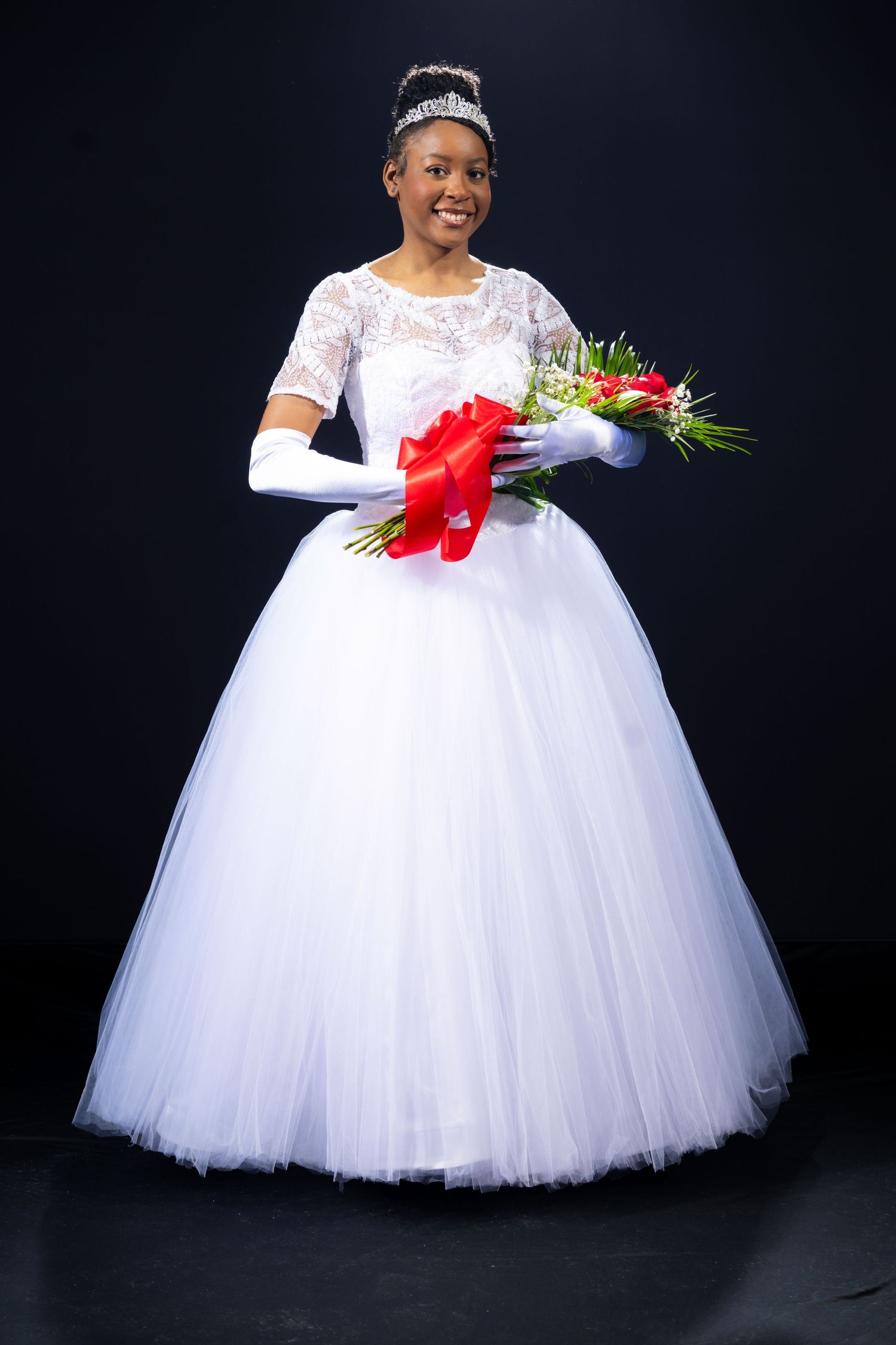Young woman in white ball gown with tiara and gloves holds flower bouquet with red bow against black background.