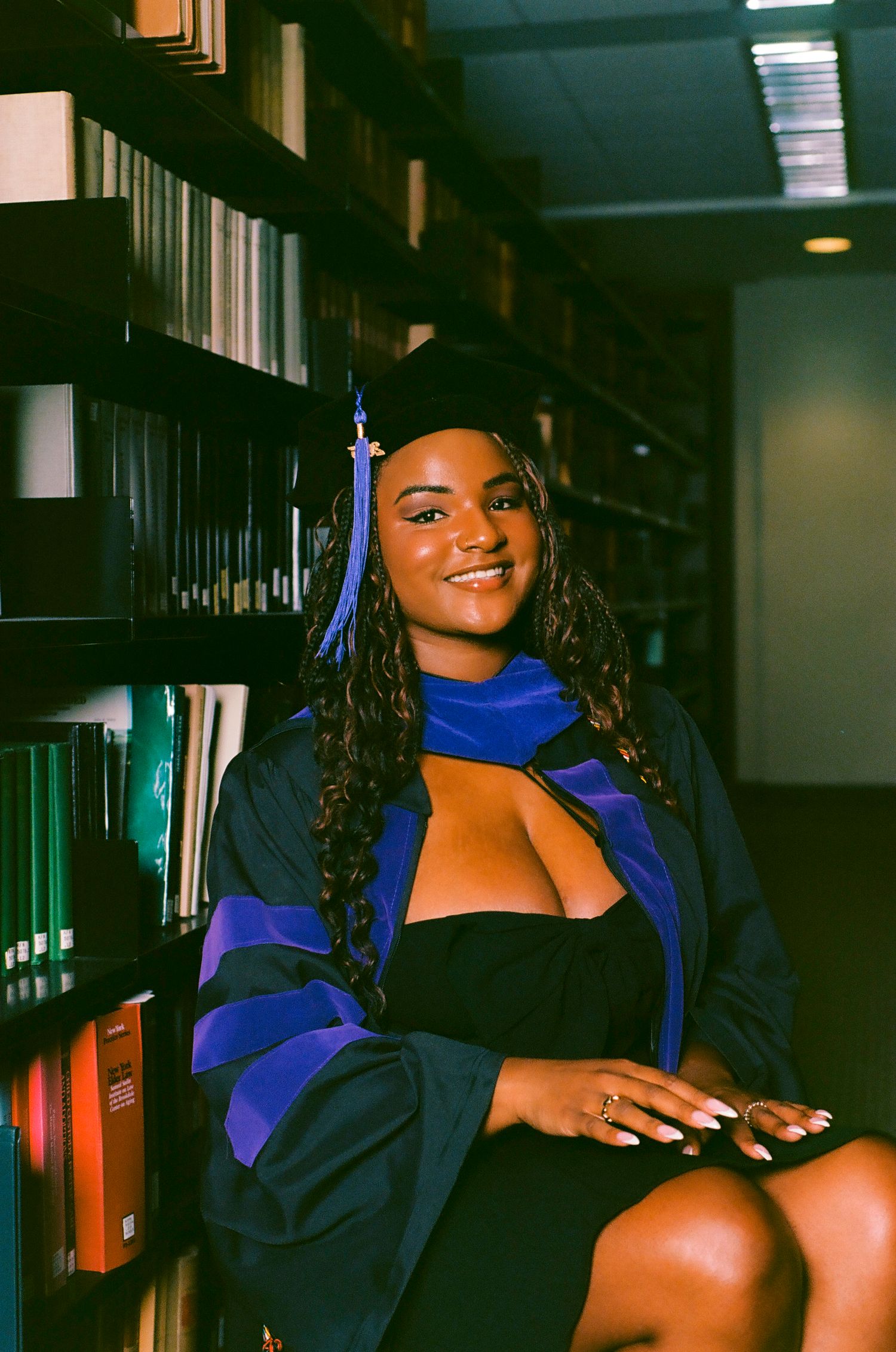 A smiling graduate in cap and gown poses beside library bookshelves in blue and black academic regalia.