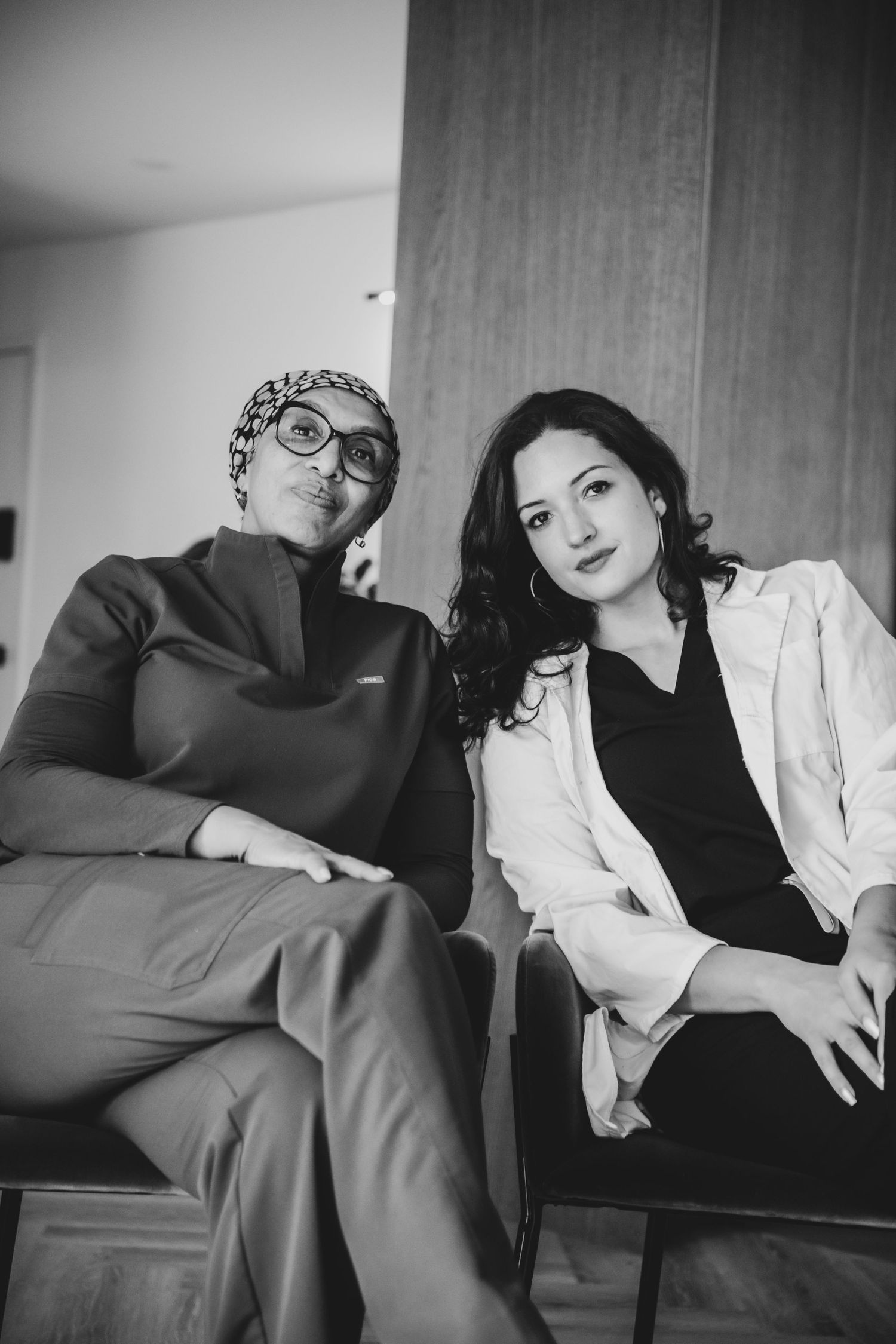 Black and white photo of two women sitting together, one wearing glasses and a head covering, the other in a white blazer.