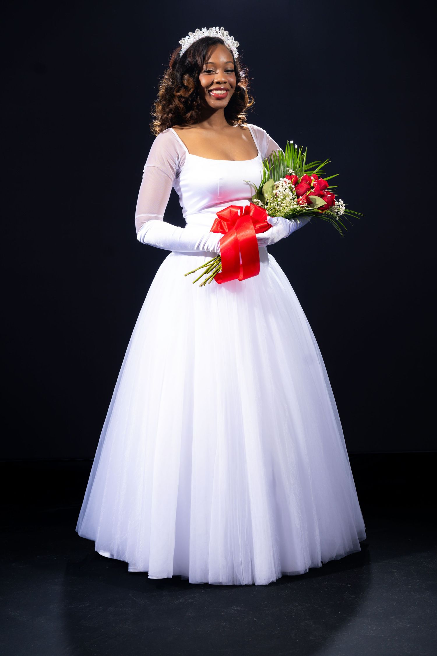 Smiling Black bride in white ball gown with sheer gloves, floral crown, holding red-ribboned bouquet against dark background.