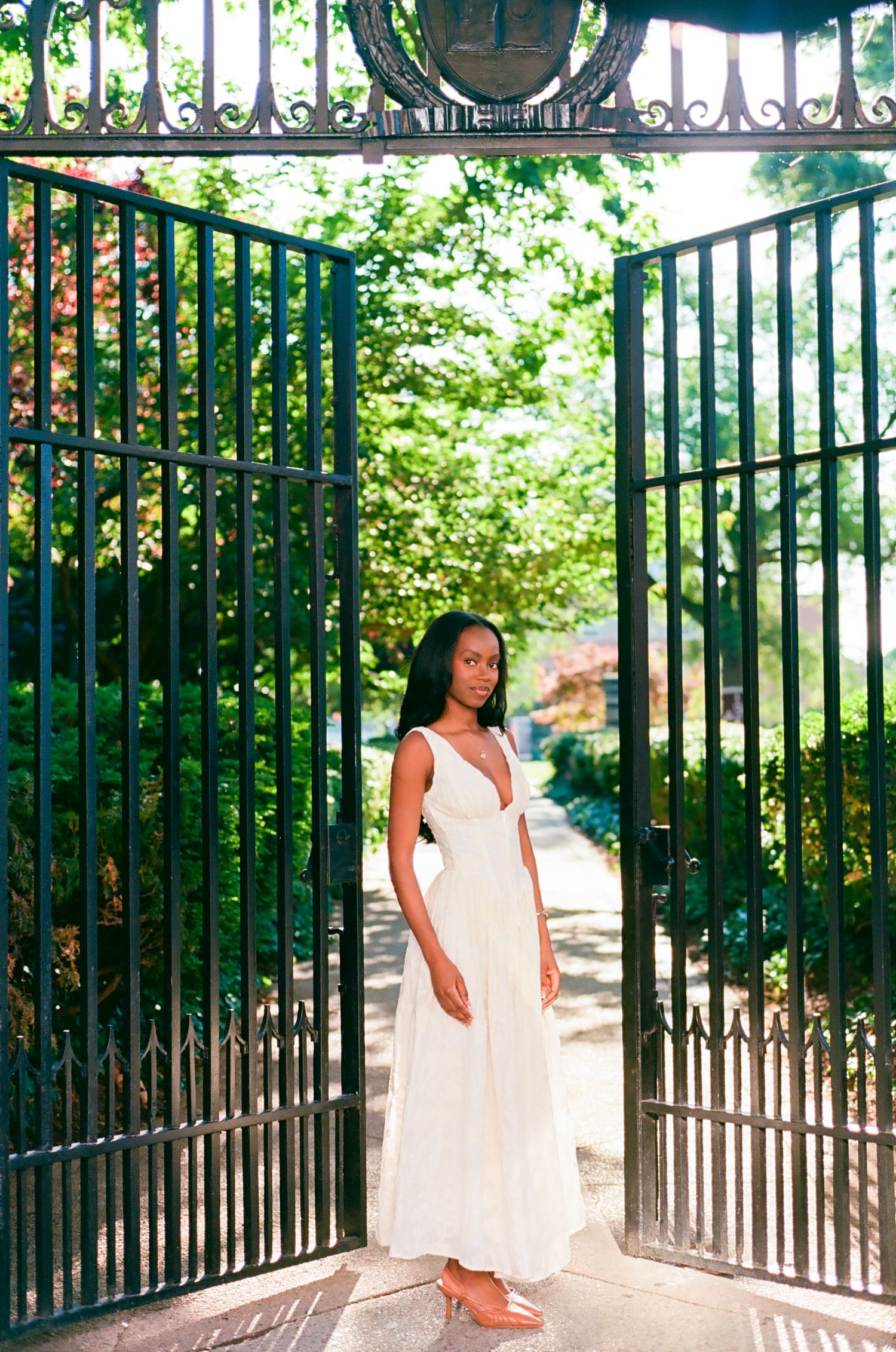 A young Black woman in a white maxi dress stands gracefully between ornate iron gates surrounded by lush greenery.