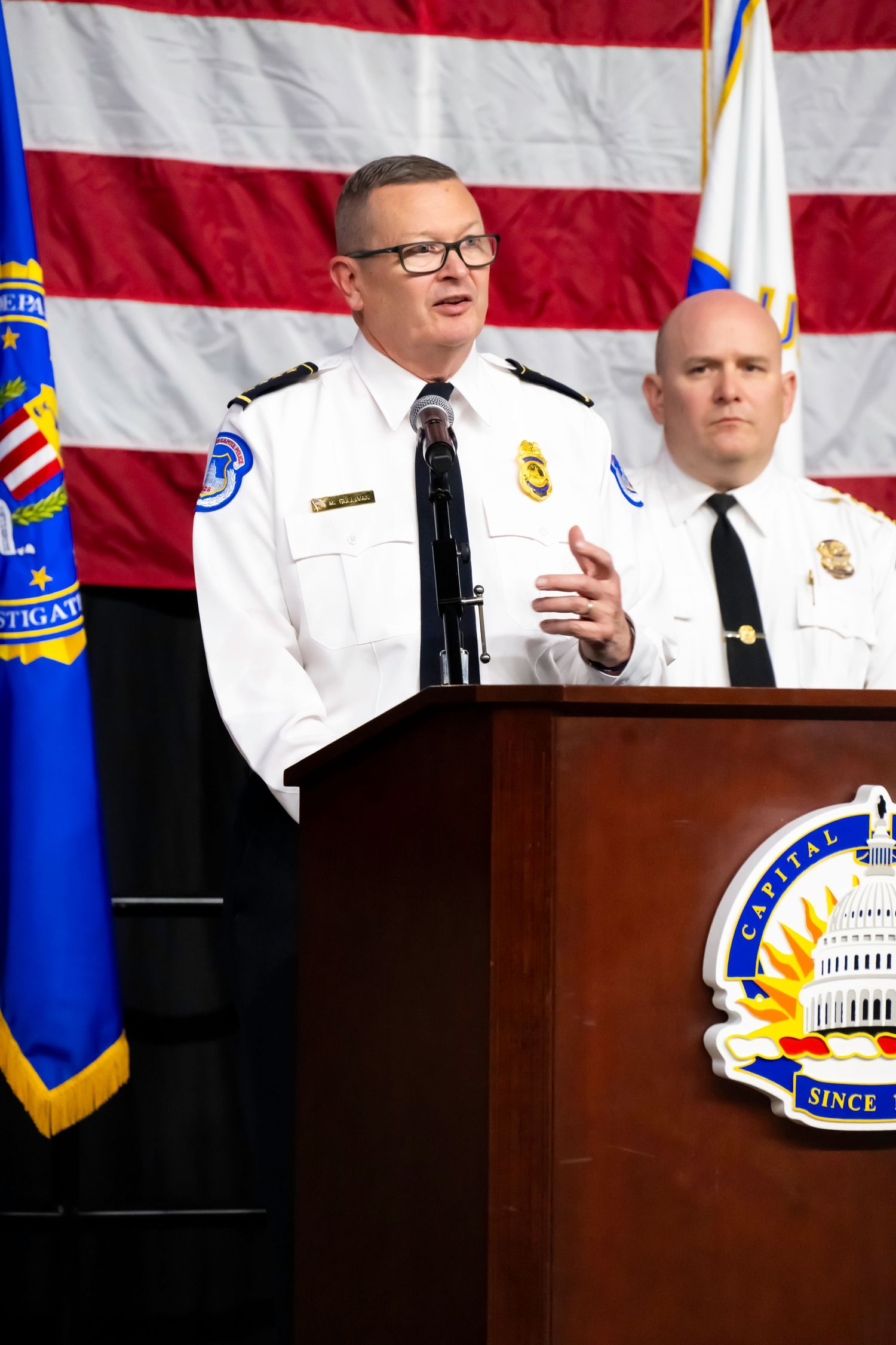 Police chief speaks at podium with U.S. flag backdrop during official law enforcement press conference.