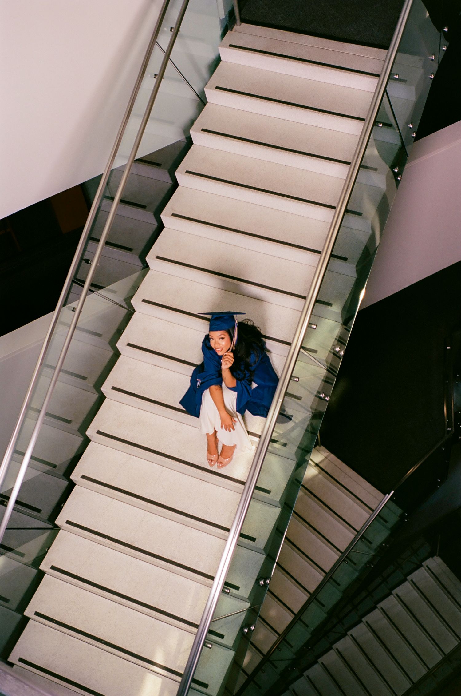 Graduate in blue cap and gown sits on modern white staircase, photographed from above.