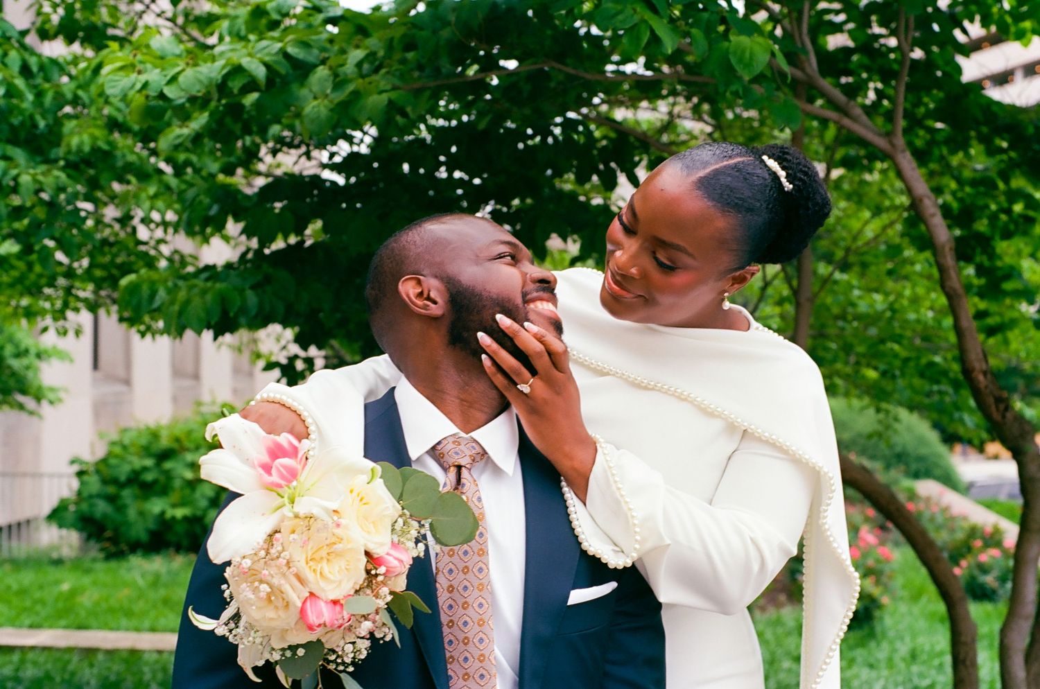 A bride in a white cape gown tenderly touches her groom's face as he holds a pink and white bouquet outdoors.