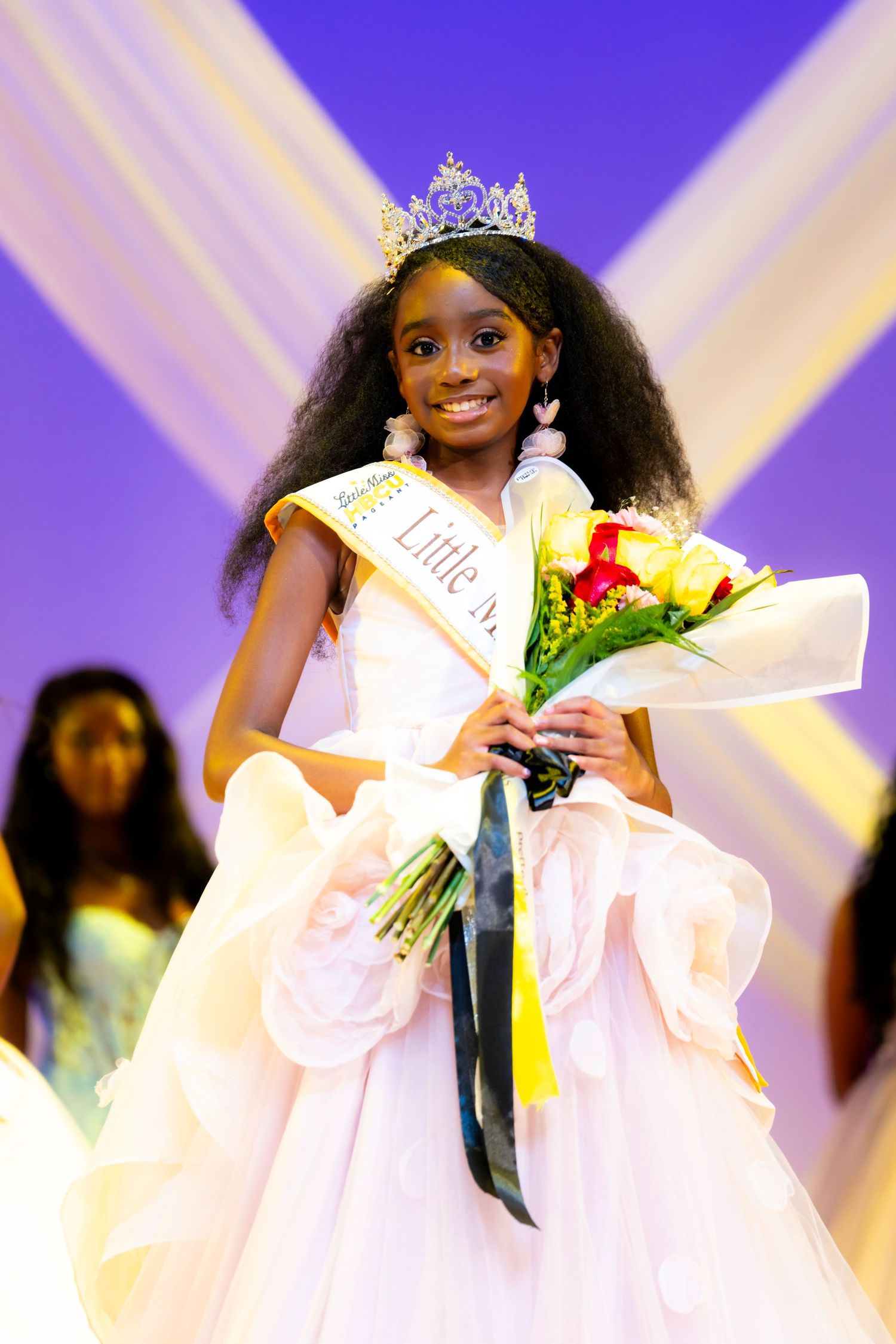 Young girl in white gown, tiara, and sash holds bouquet, smiling as a beauty pageant winner on stage.