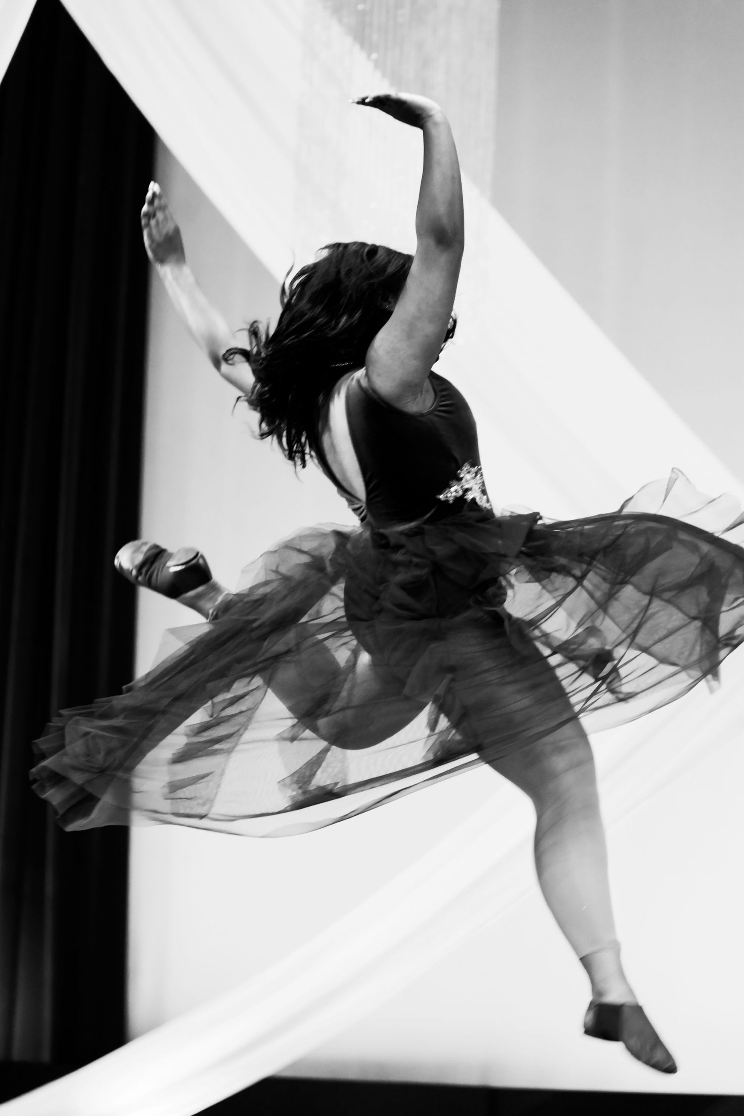 Black and white photo of a ballerina mid-leap in a tutu, arms raised, captured with motion blur on stage.
