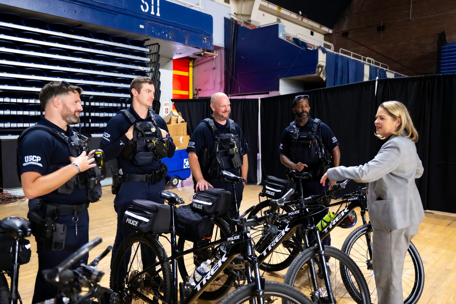 Four police officers in tactical gear chat with a woman near bicycles inside a large indoor venue.