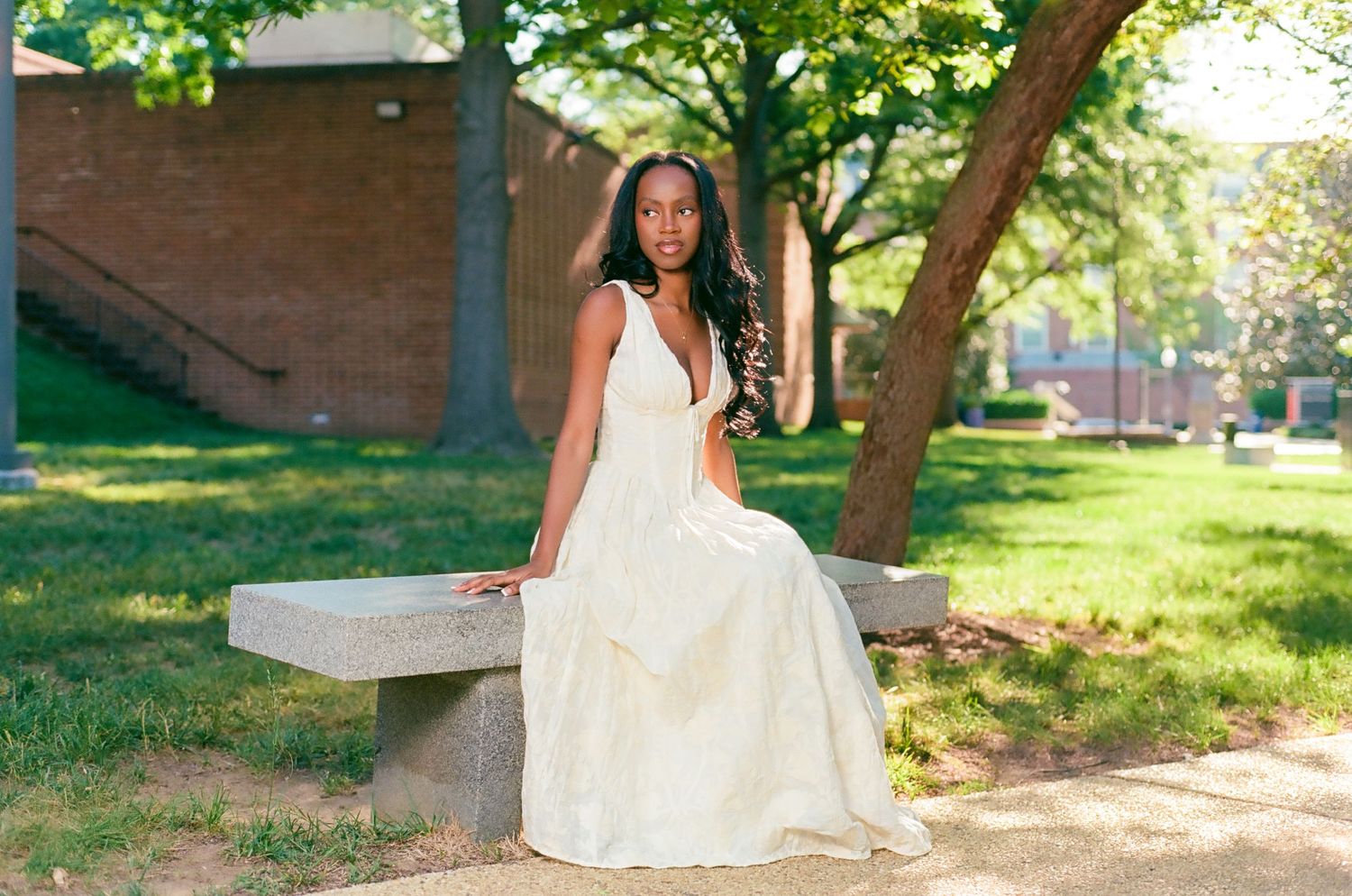 A young Black woman in an elegant white gown sits gracefully on a stone bench in a sunlit park.