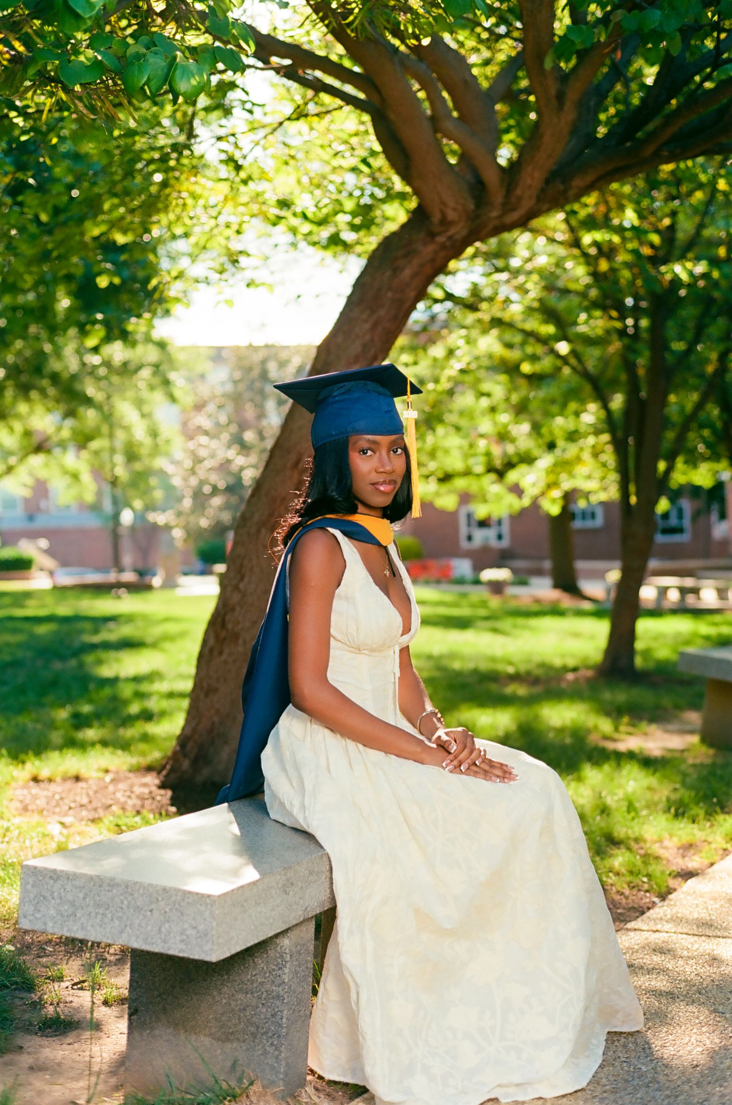 A young woman in a white dress and navy graduation cap and gown sits on a stone bench under a tree.