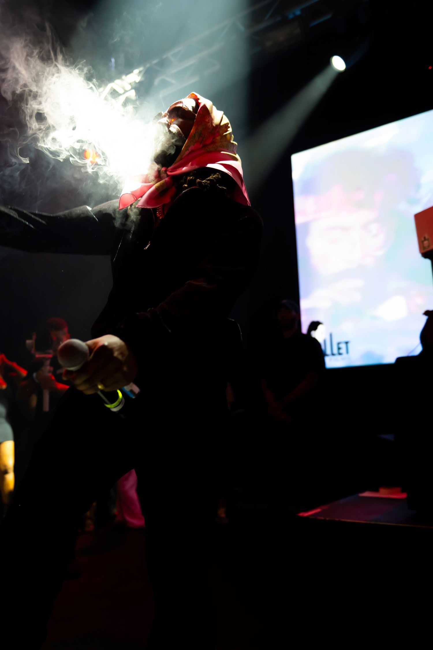 Rapper performing on stage under dramatic spotlights with smoke effects and a screen display in the background.
