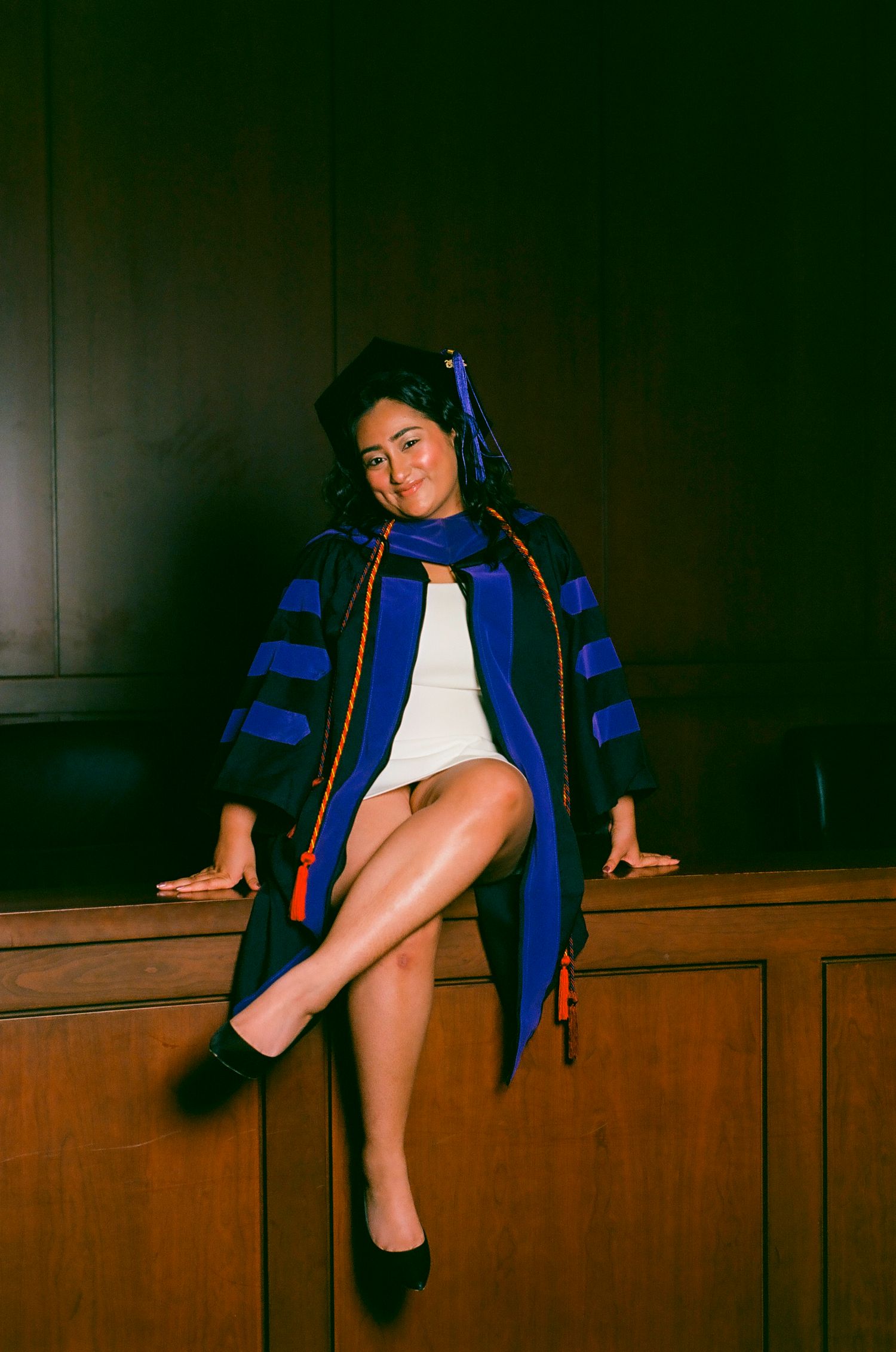 Woman in blue doctoral graduation regalia and cap sits confidently on wooden furniture, posing for a celebratory photo.