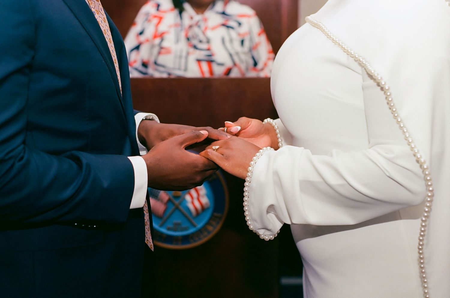 Couple exchanging wedding rings during a church ceremony, groom in blue suit, bride in white dress with pearl accessories.