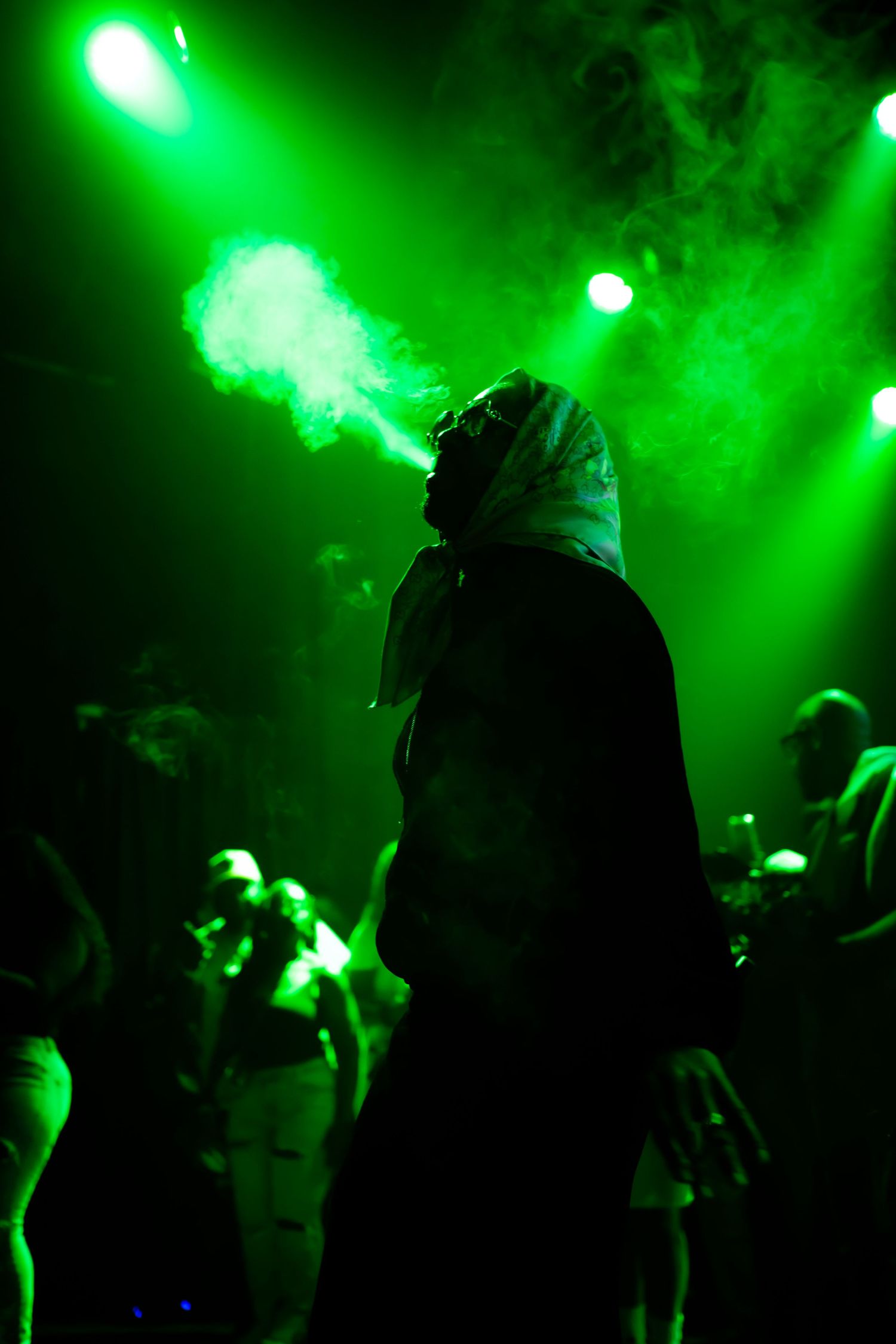 Silhouetted performer exhaling smoke on stage under dramatic green concert lights with crowd in background.
