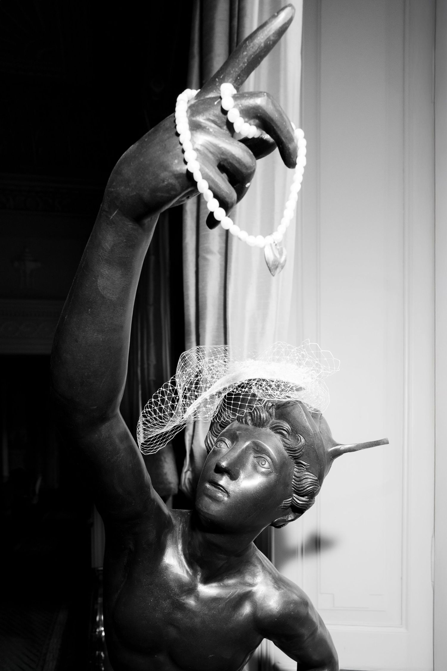 Black and white photograph of a classical bronze statue holding up a pearl necklace in an artistic pose.