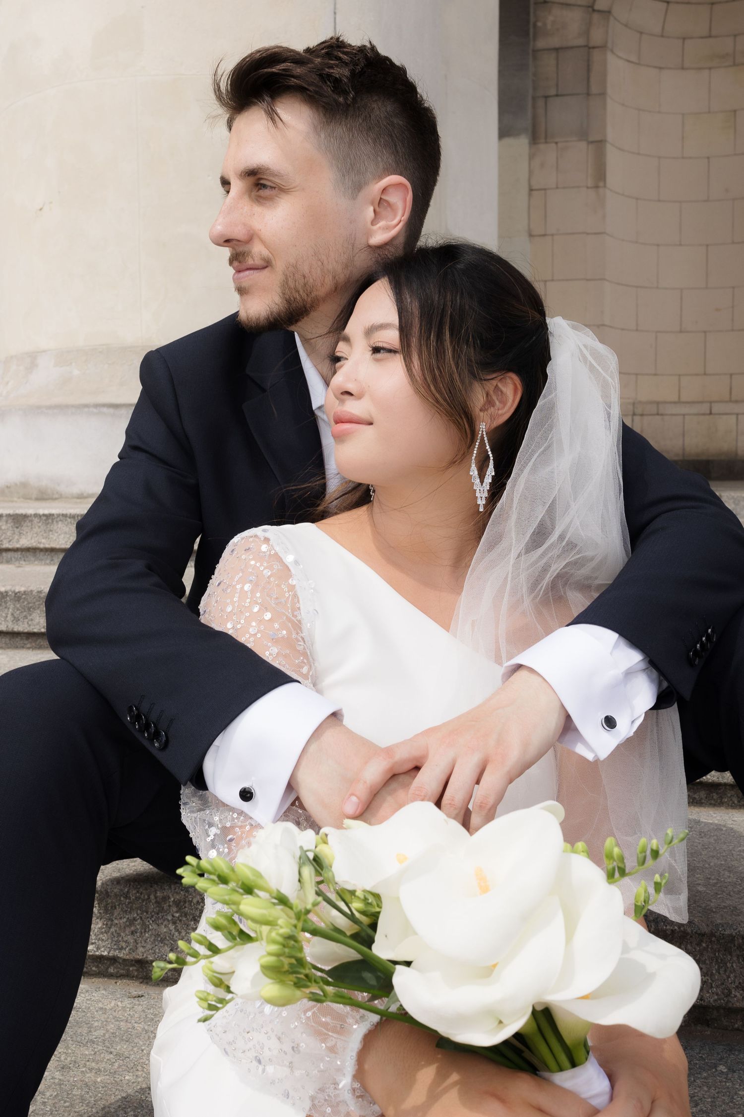 A couple in wedding attire embraces while holding white floral bouquet.