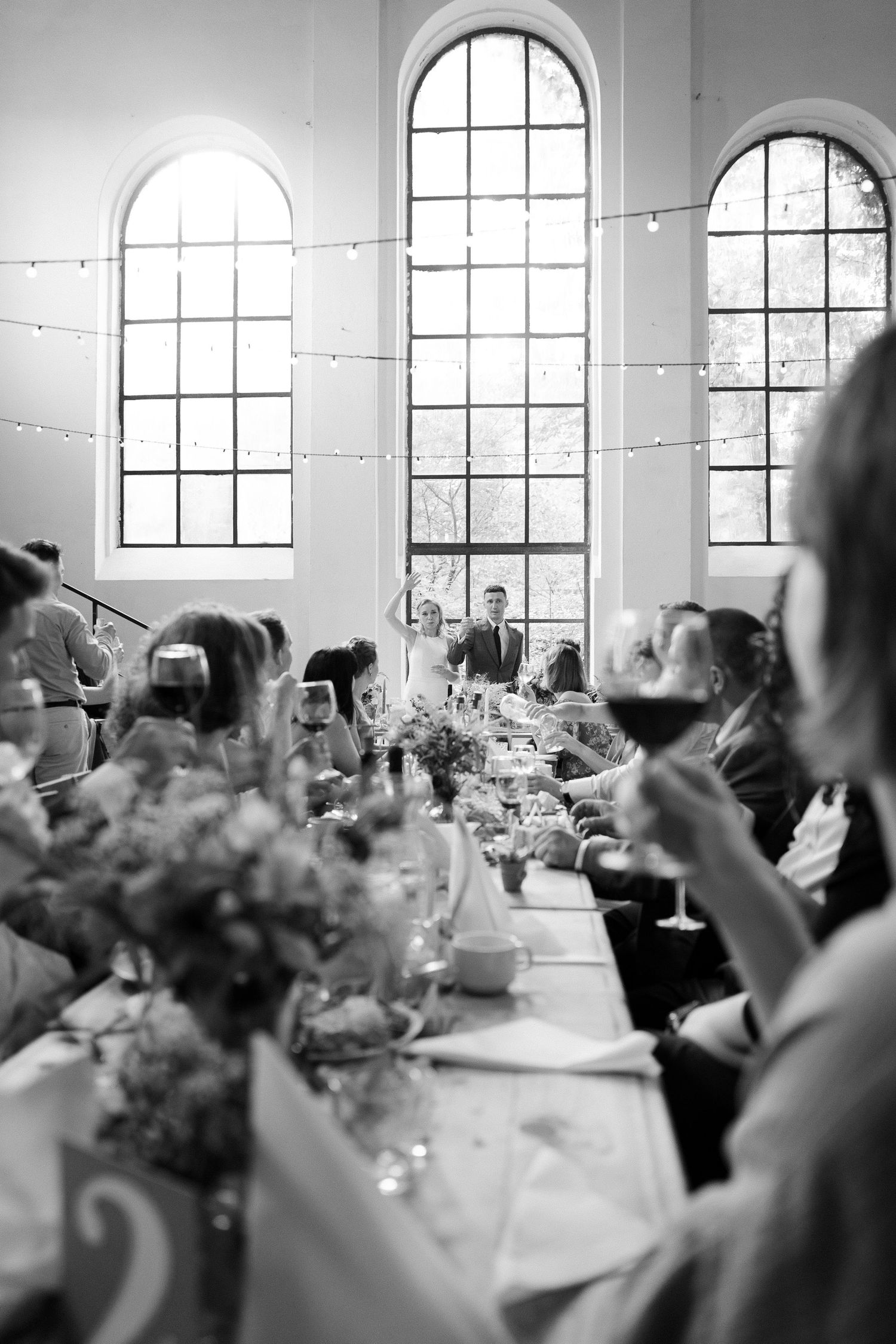 Black and white panoramic view of an elegant wedding reception venue with arched windows and tables set for dining.