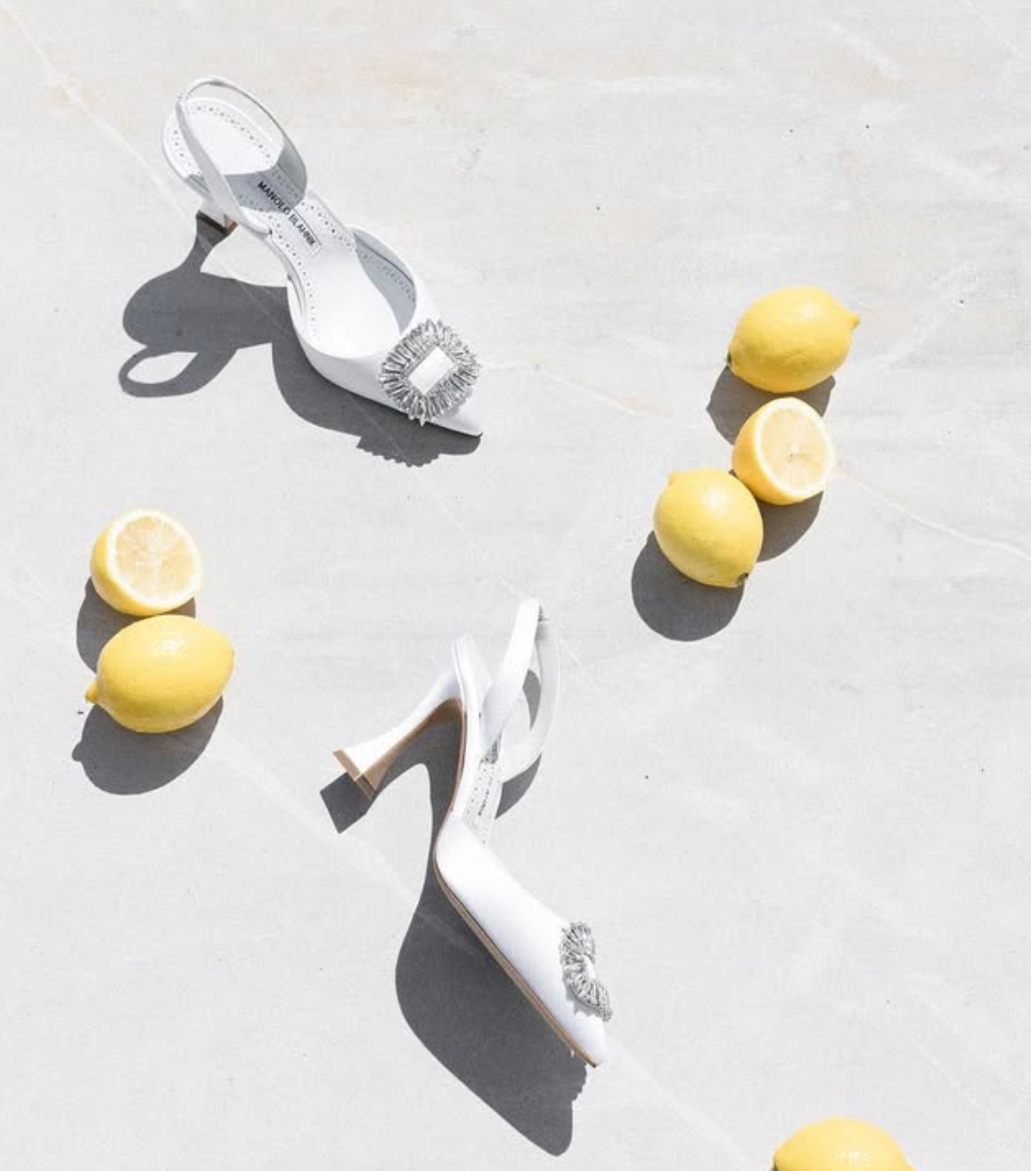 White high heel sandals and fresh lemons arranged on light background with dramatic shadows.
